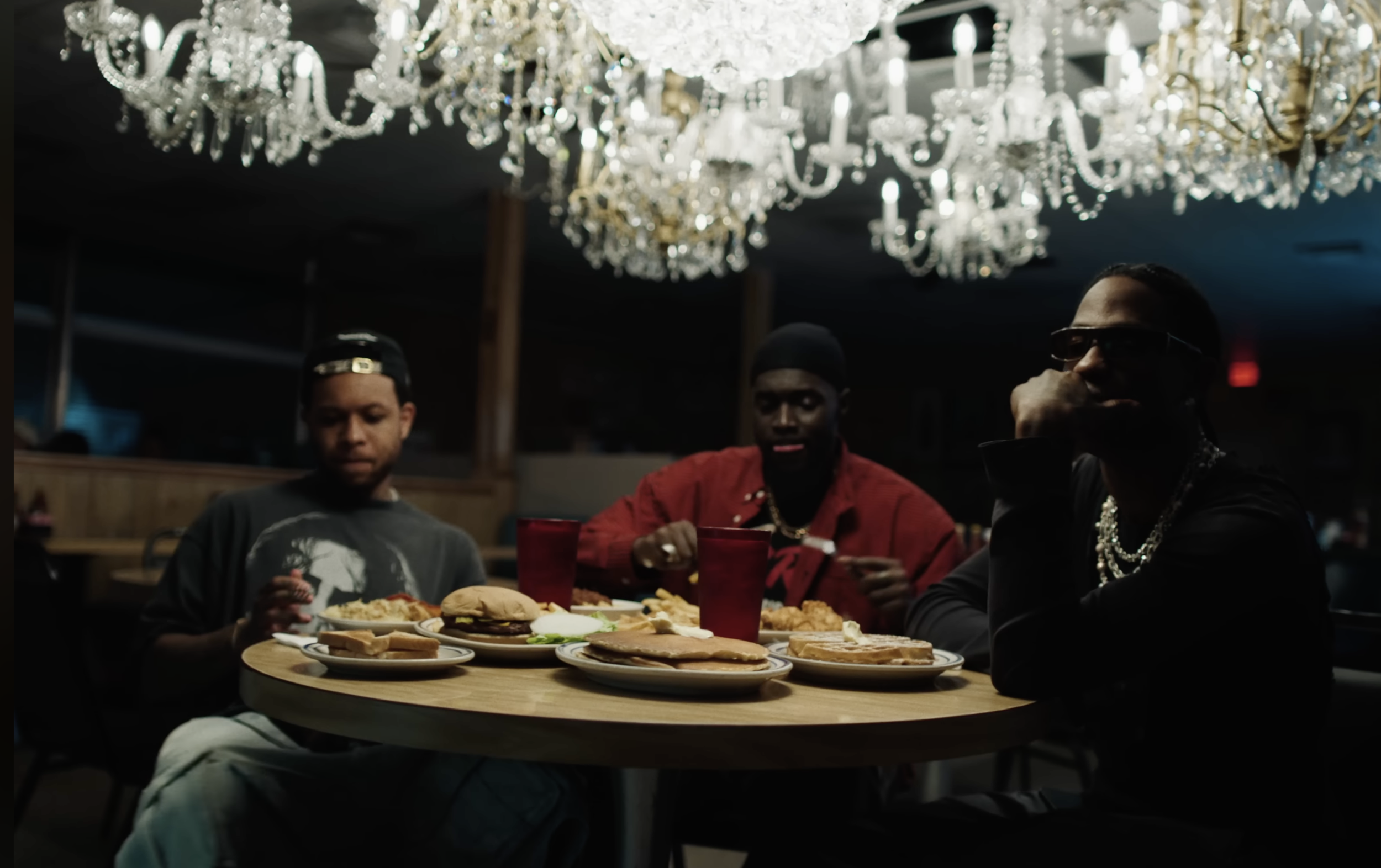 Three young men sitting at a table in a dimly lit restaurant with a chandelier hanging above. They are eating and have various plates of food, including sandwiches and fried items. Two of the men are wearing dark clothing, and one is wearing a red ja