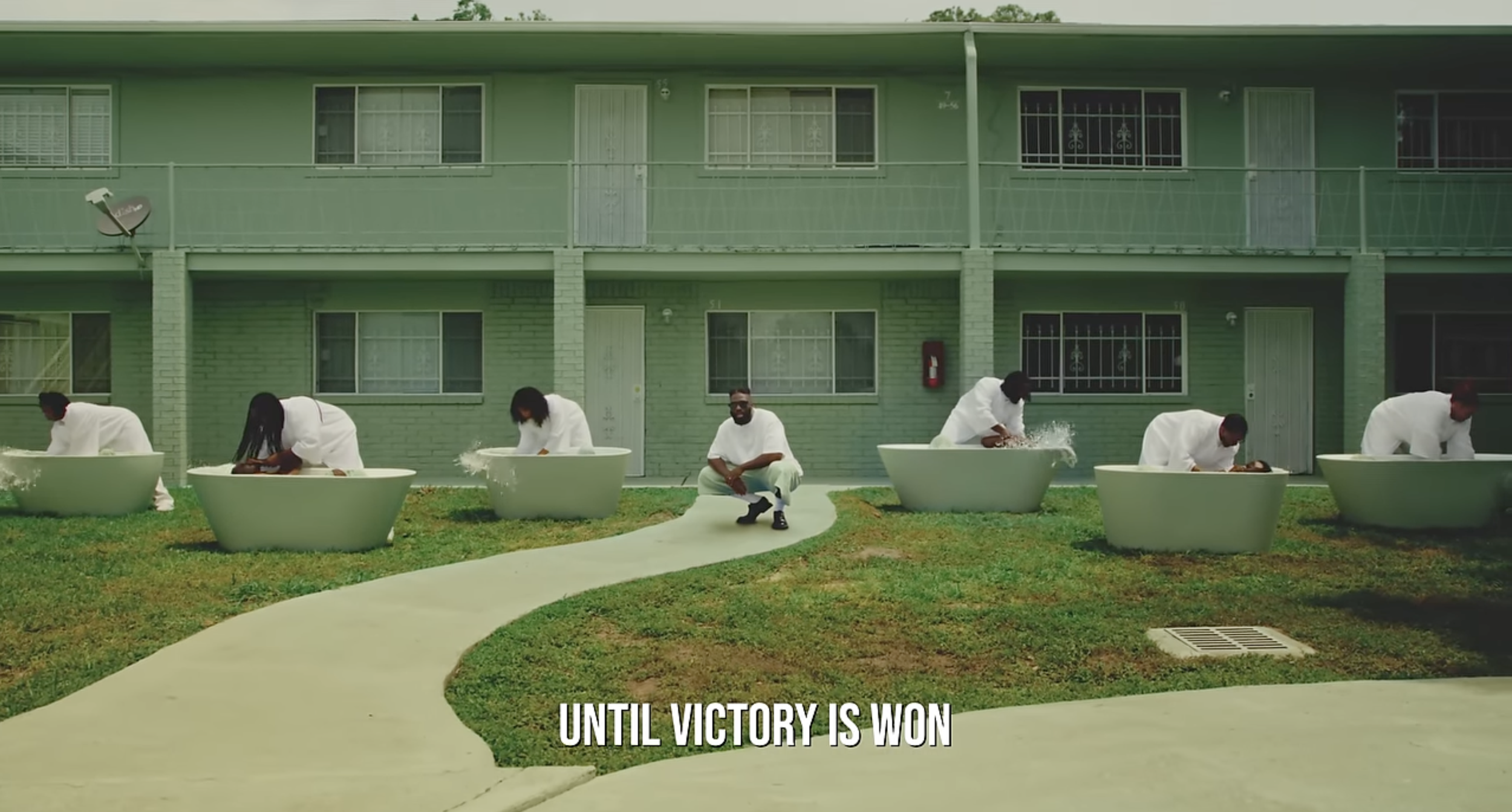 Six people dressed in white clothing positioned around large white bathtubs outside a green apartment building, with one person sitting on a sidewalk. The scene shows a grassy area with a winding walkway and a drainage grate in the foreground. Captio
