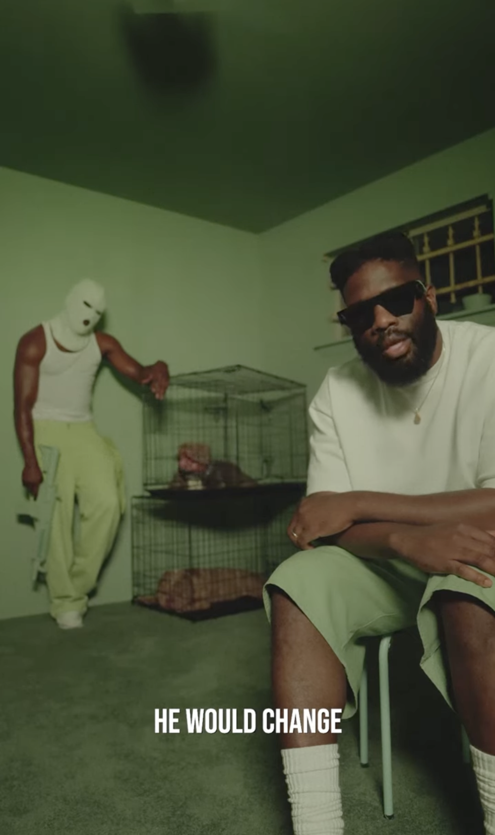 A man with sunglasses sitting on a chair in a room painted green, with a person in a white mask and yellow pants standing beside a cage with animals inside. The text overlay reads, "HE WOULD CHANGE."
