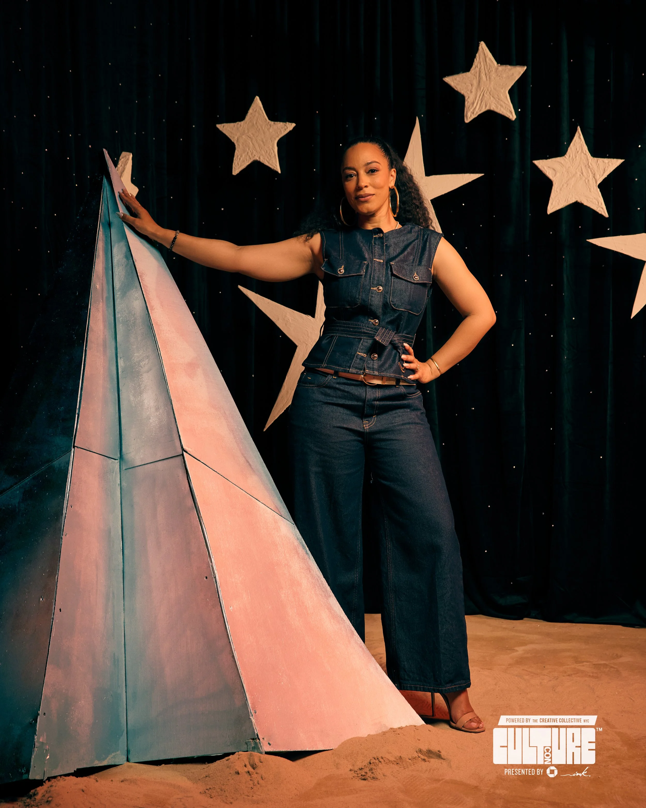 A woman in denim outfit standing next to a painted triangular sculpture on stage with black curtain and large star decorations in background.