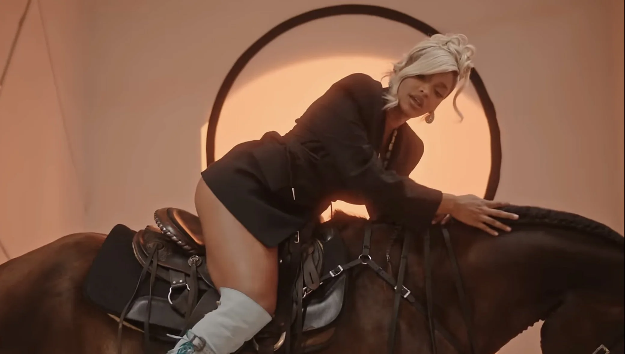 Woman with blonde hair riding a brown horse, wearing a black jacket and riding gear, indoors with a glowing circular background.