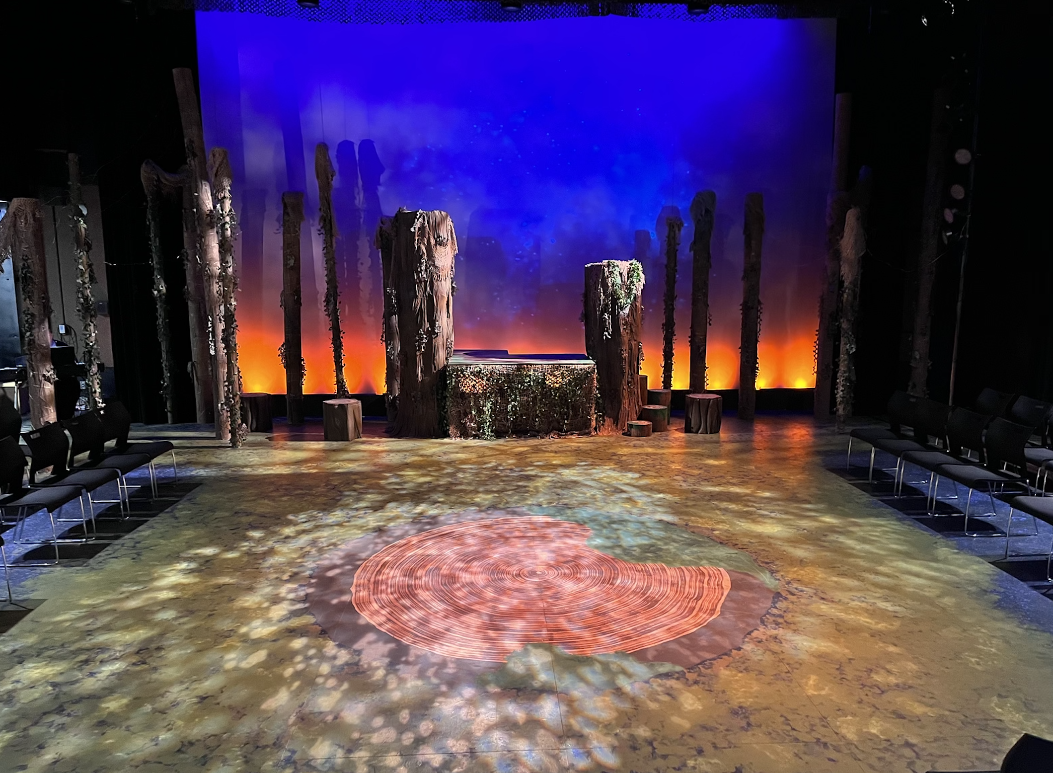 Stage set up for a theatrical performance with tall wooden pillars decorated with moss and vines, a backdrop of a starry night sky with clouds, warm orange lighting at the base of the pillars, and a textured floor with a circular light projection res