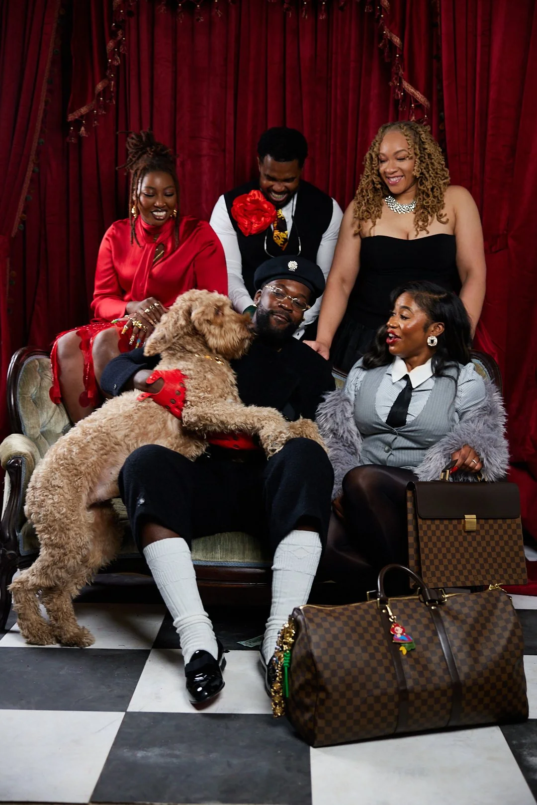 A group of six people and a dog, seated and standing in front of red curtains, with bags on the floor, posing for a photo.