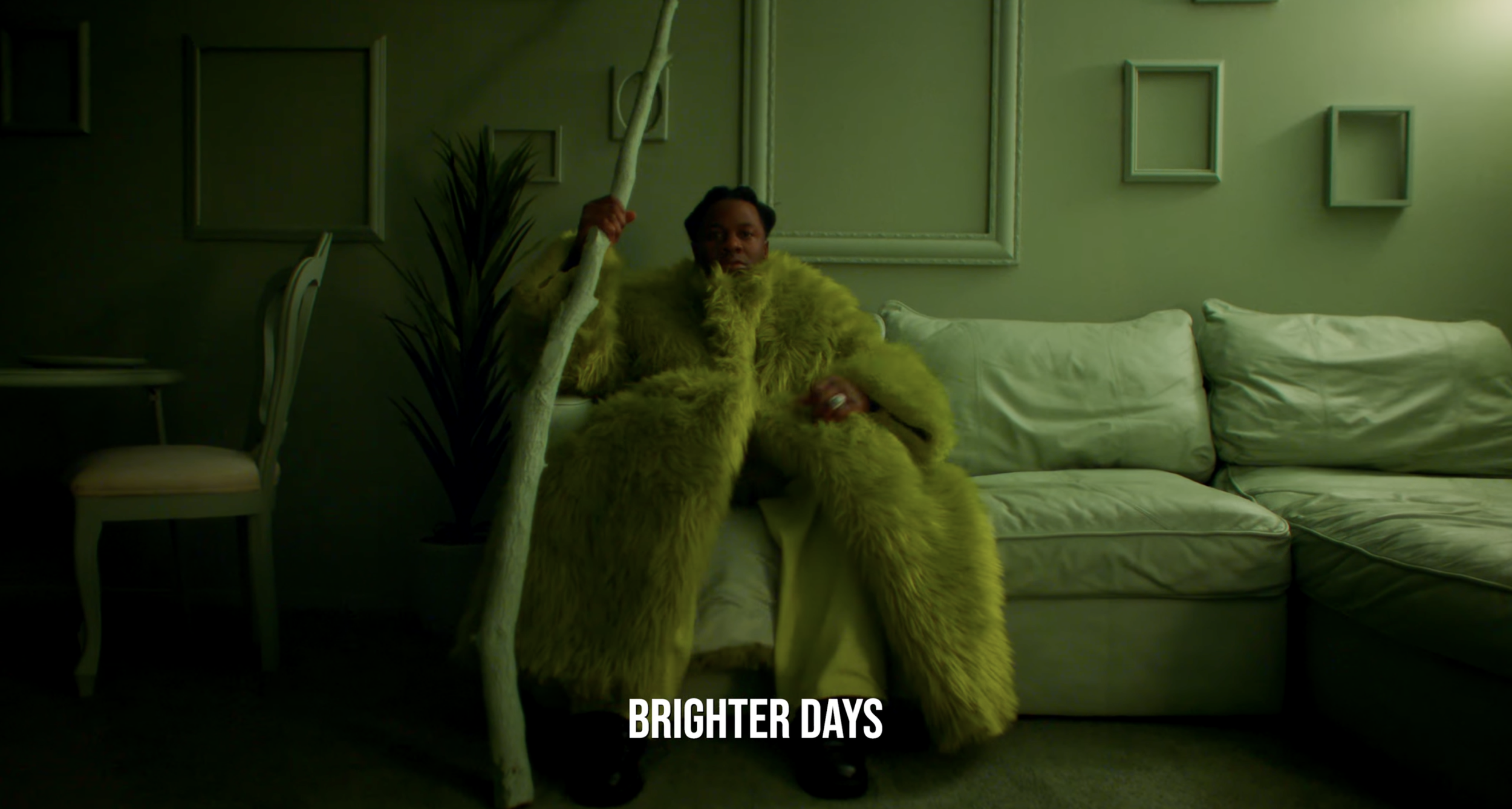 A person sitting on a white couch holding a tall white staff, wearing a bright yellow furry coat. There are empty picture frames on the green wall behind and a potted plant nearby. The image has the text 'Brighter Days' at the bottom.