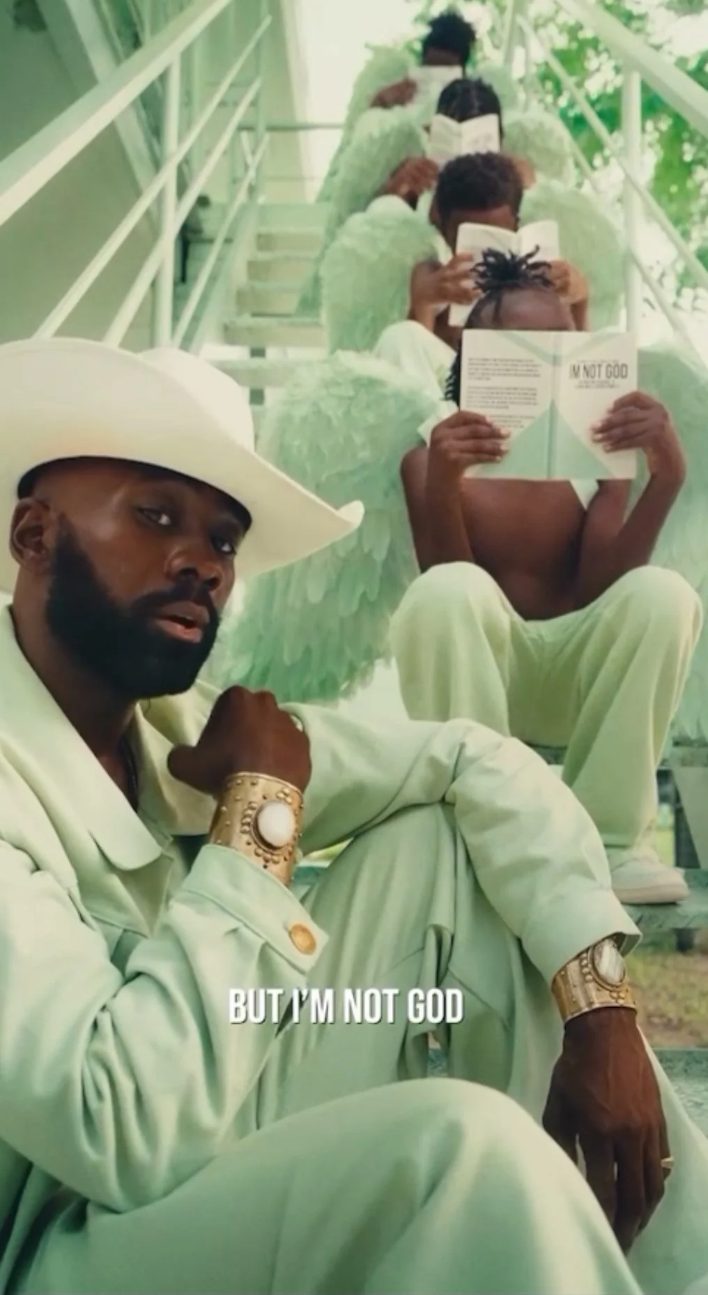 A man dressed in a light green suit and wide-brimmed hat sitting on stairs with six people behind him, all wearing similar outfits and reading books, with green angel wings painted on their backs. The man in the foreground is looking at the camera an