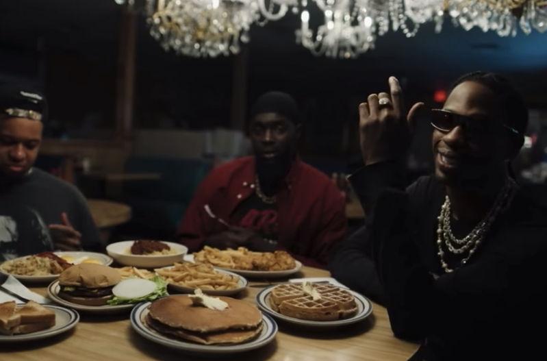 Three men sitting at a table with various food dishes in a dimly lit restaurant, decorated with chandeliers overhead.