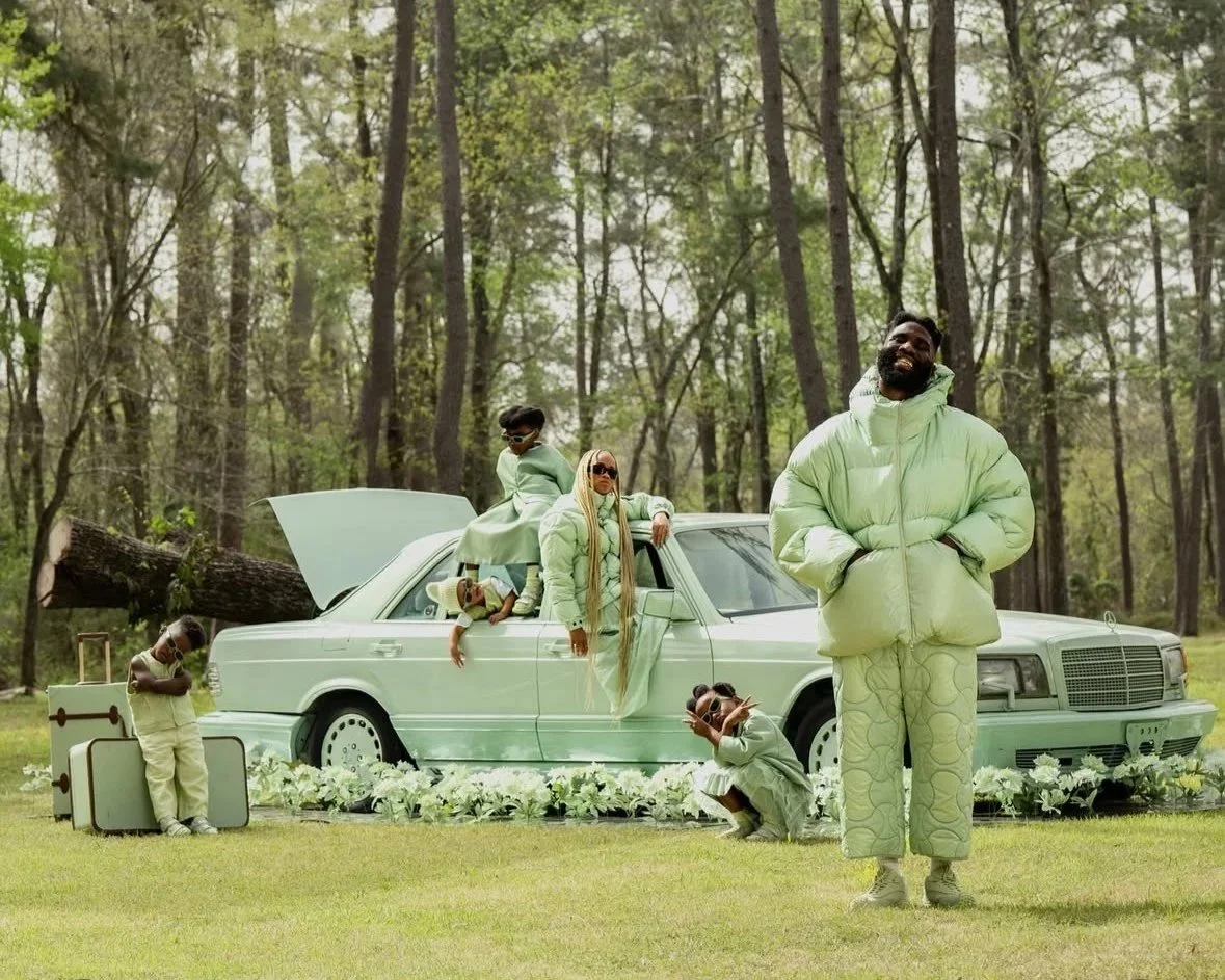 A group of people dressed in coordinated light green clothing poses outdoors around a white vintage car, with some sitting and standing on and around it, in a forest setting.