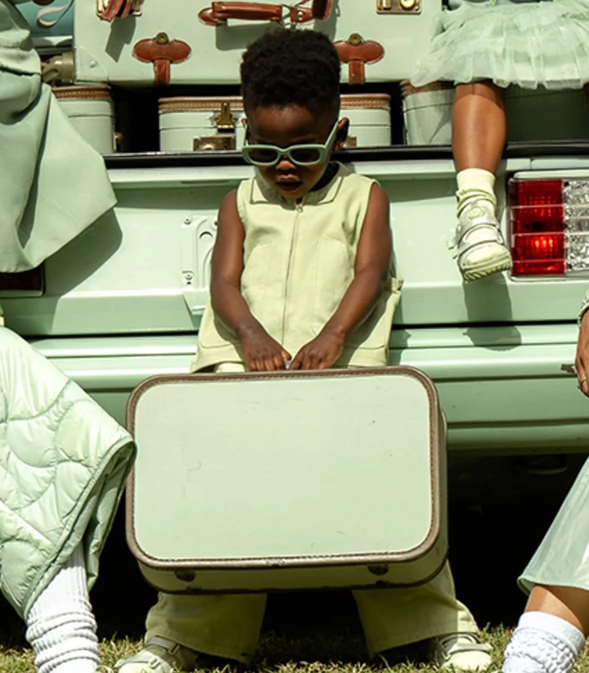 A young boy wearing sunglasses and a beige vest standing behind a beige suitcase at an outdoor yard sale or flea market. There are other items and furniture visible in the background.