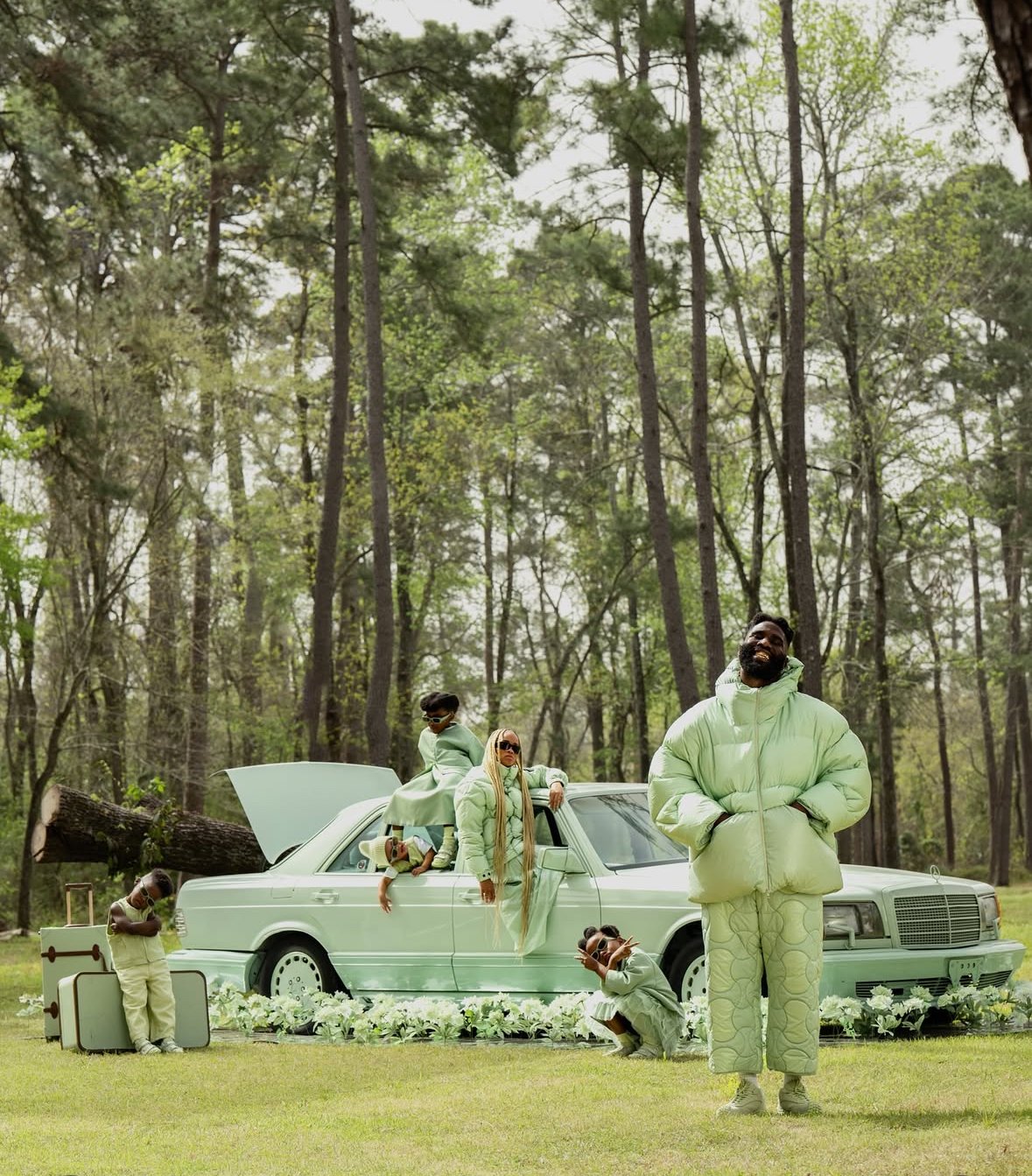 A group of six people with dark skin posing outdoors in front of a white vintage car in a forest setting, all dressed in light green clothing. The scene includes floral decorations and a suitcase. The individuals are striking various poses, with some