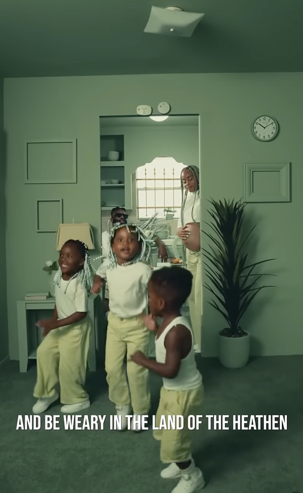 A woman and four children dancing in a living room with green walls, a clock, and houseplants, while singing the lyrics 'And be weary in the land of the heathen.'