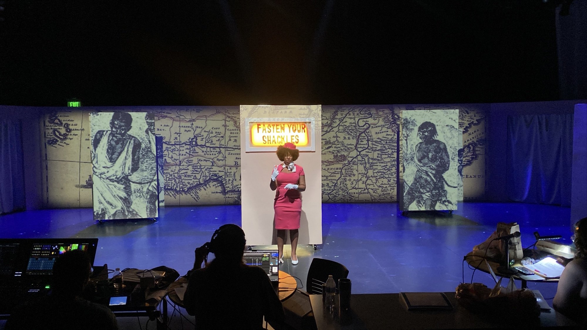 Theatre stage with a woman in a pink dress and hat standing in front of a backdrop with old maps and black-and-white photos, and a neon sign that says "FASTEN YOUR SHACKLES." Troupes of people are in the foreground, including a person working control