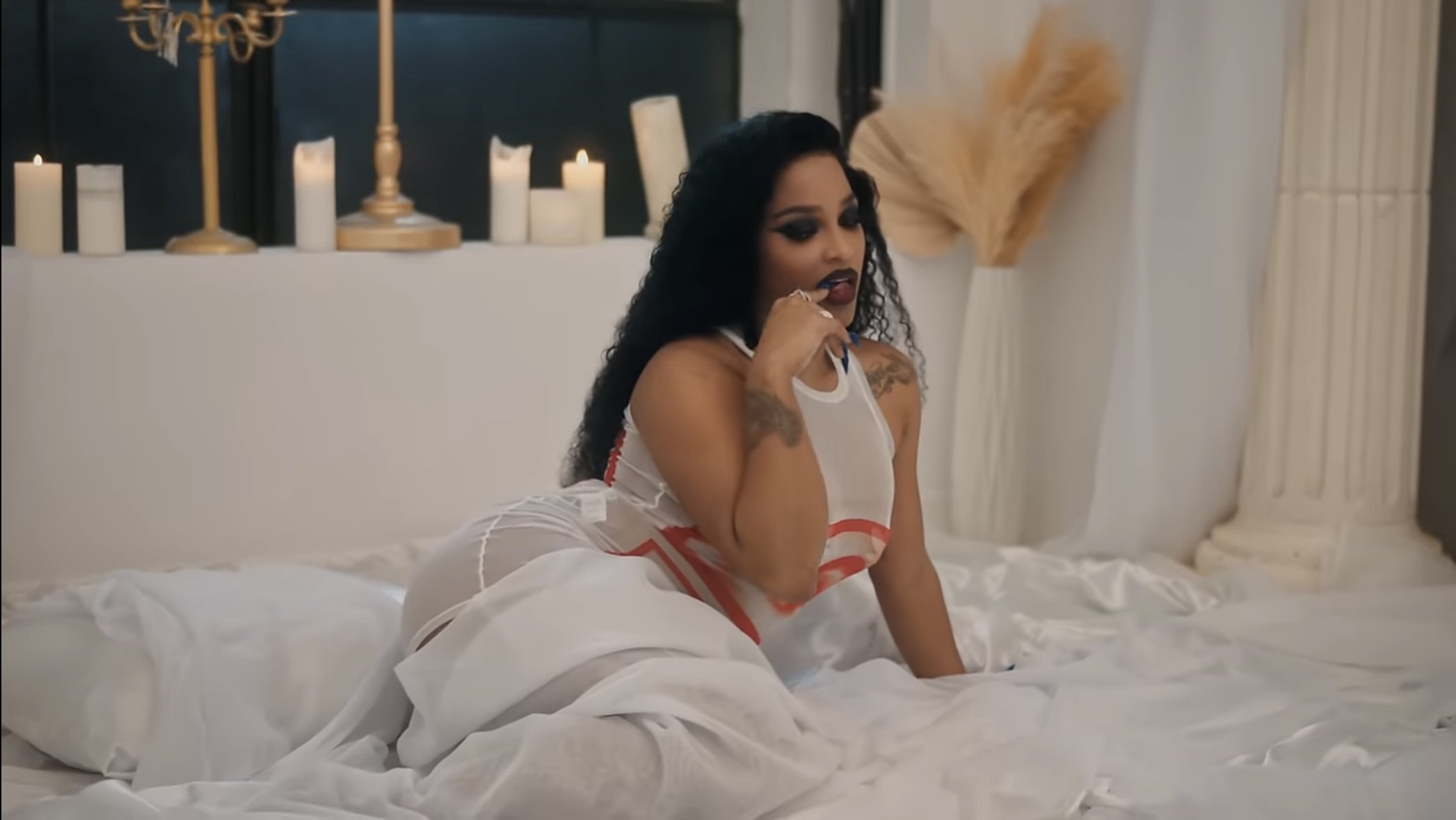 A woman with dark hair and tattoos on her arms is posing on a bed with white linens, wearing a sheer white and red outfit, in a room with candles and decorative elements.
