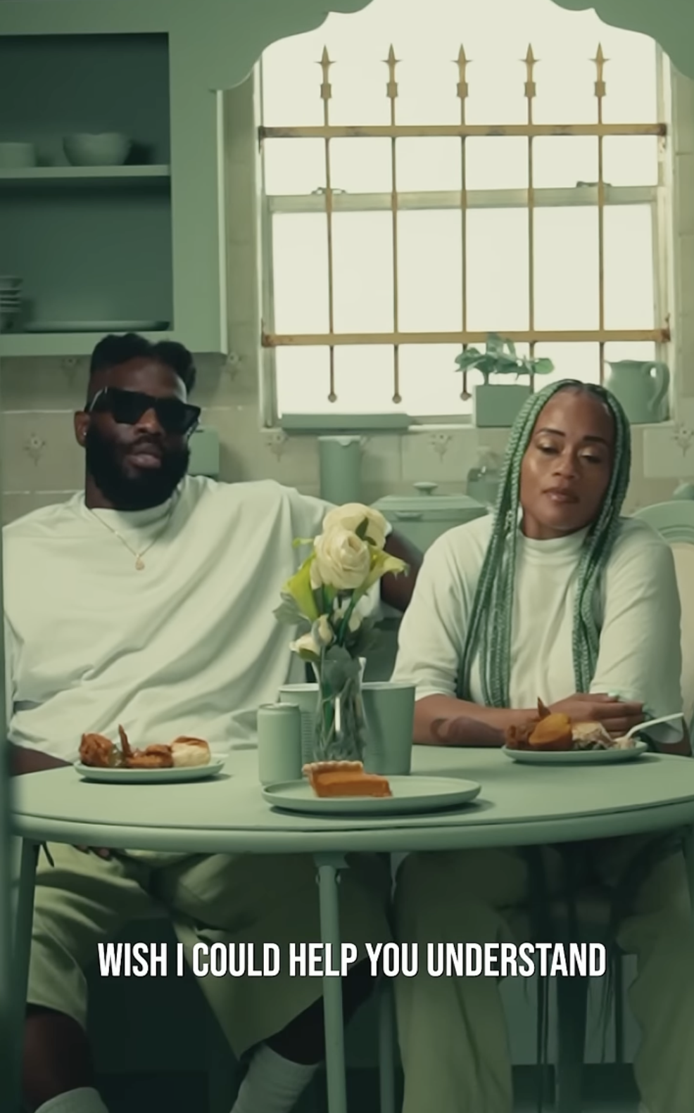 Two people sitting at a dining table in a kitchen, with food and drinks, a flower vase, and a window with bars in the background. One person is wearing sunglasses and the other has green braided hair. The caption says, "Wish I could help you understa