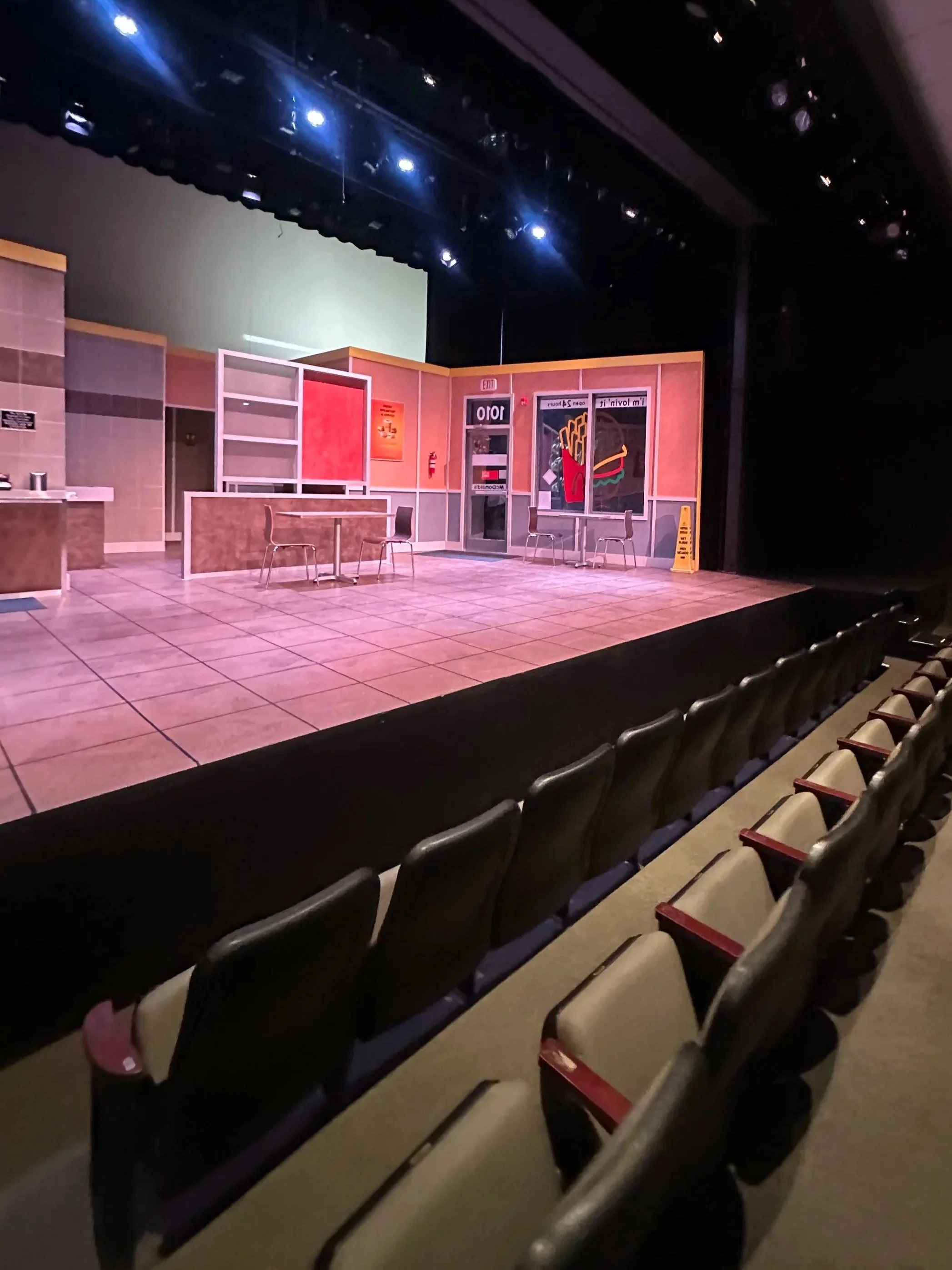 Empty theater stage with set resembling a fast food restaurant, beige chairs and tiled floor audience seats in the foreground.