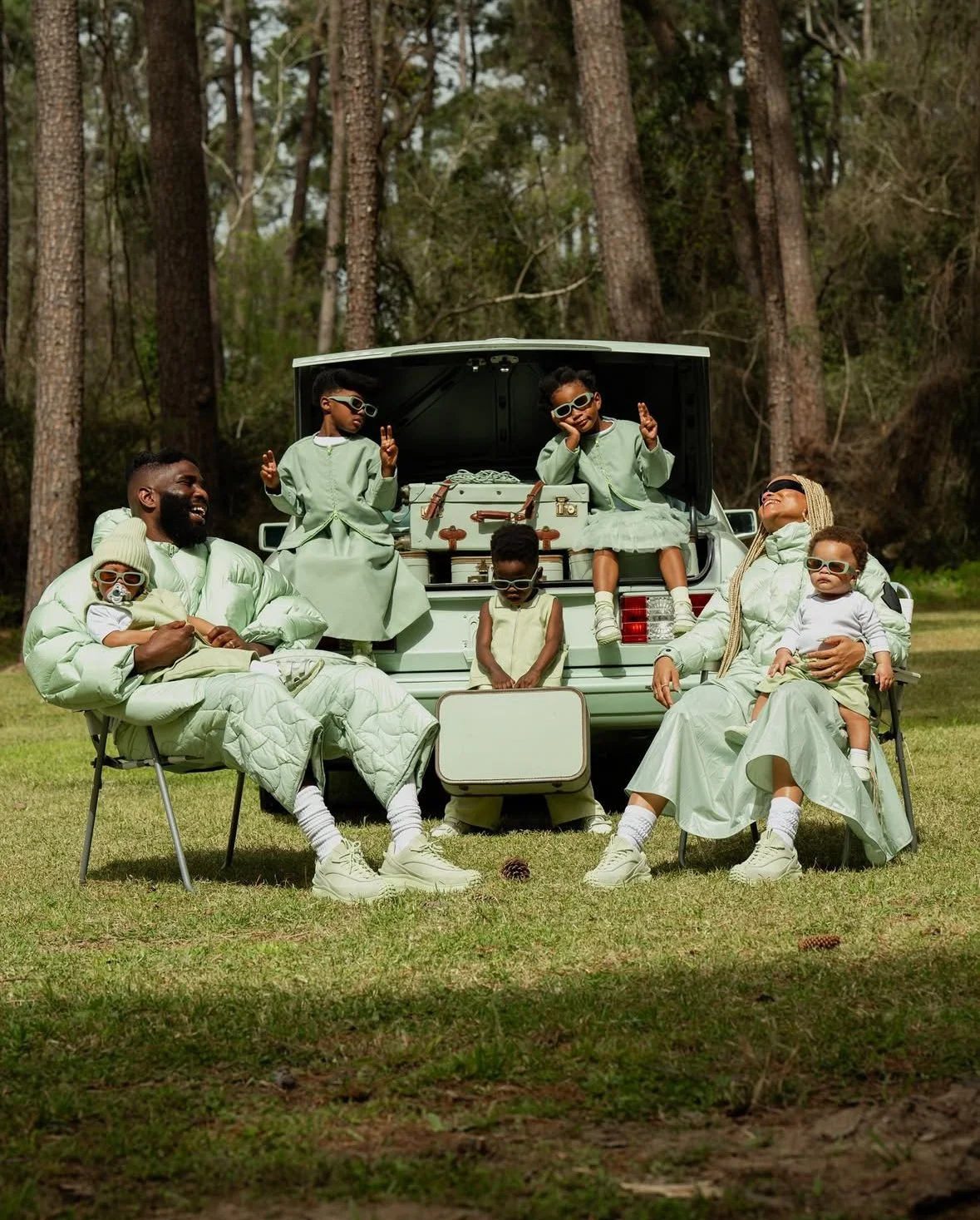 A family sitting outdoors in a forest clearing, wearing coordinated light-colored clothing, with some members wearing sunglasses. They have a vintage style picnic setup with an open car trunk behind them, filled with suitcases and picnic items. The s