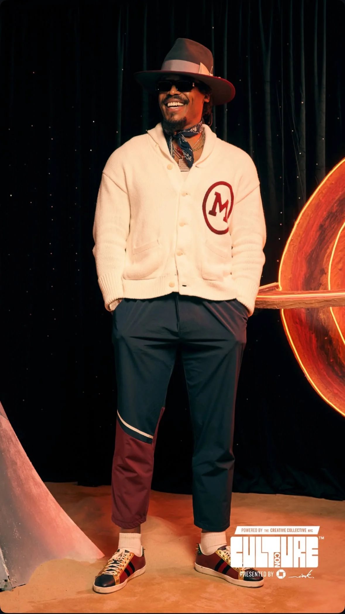 Man wearing a wide-brimmed hat, sunglasses, beige coat with an 'M' on the chest, navy pants, and sneakers, standing on a stage with a black curtain backdrop and a neon artwork.