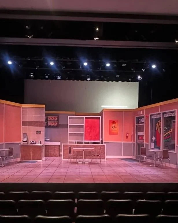 Empty theater stage set designed to look like an indoor office or service area with desks, chairs, and wall decorations.