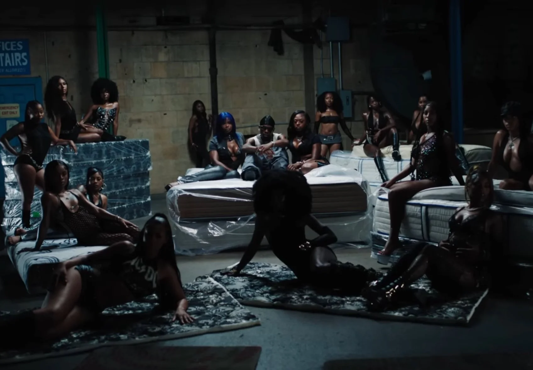 A group of women in dark clothing and shiny outfits in an industrial space with mattresses and bedding, some sitting and some lying on the floor, creating a scene that appears staged or part of a performance.