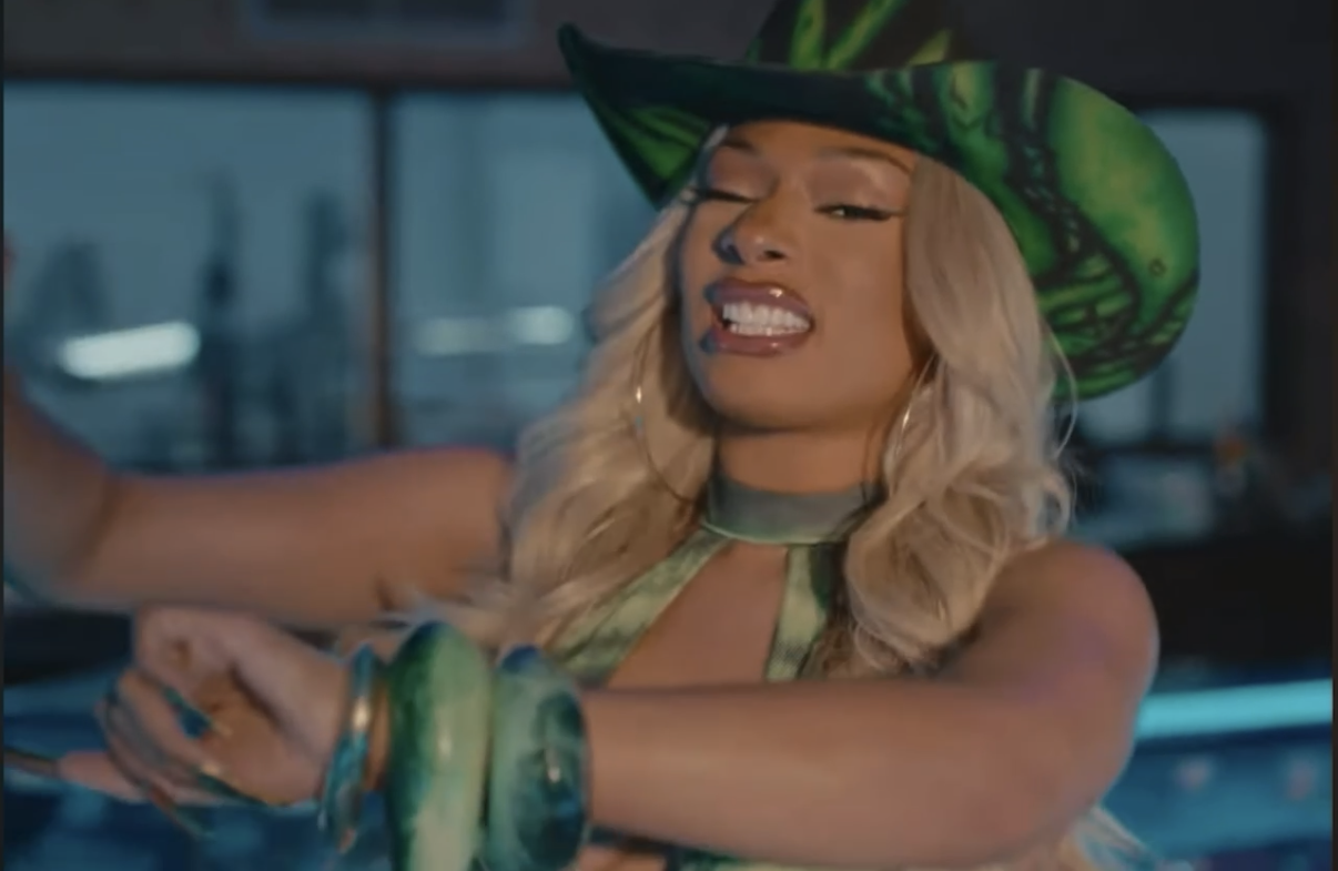A woman with blonde hair wearing a large green hat with snake pattern, a matching top, and jewelry, appears to be taking a selfie or video in a kitchen or bar setting.