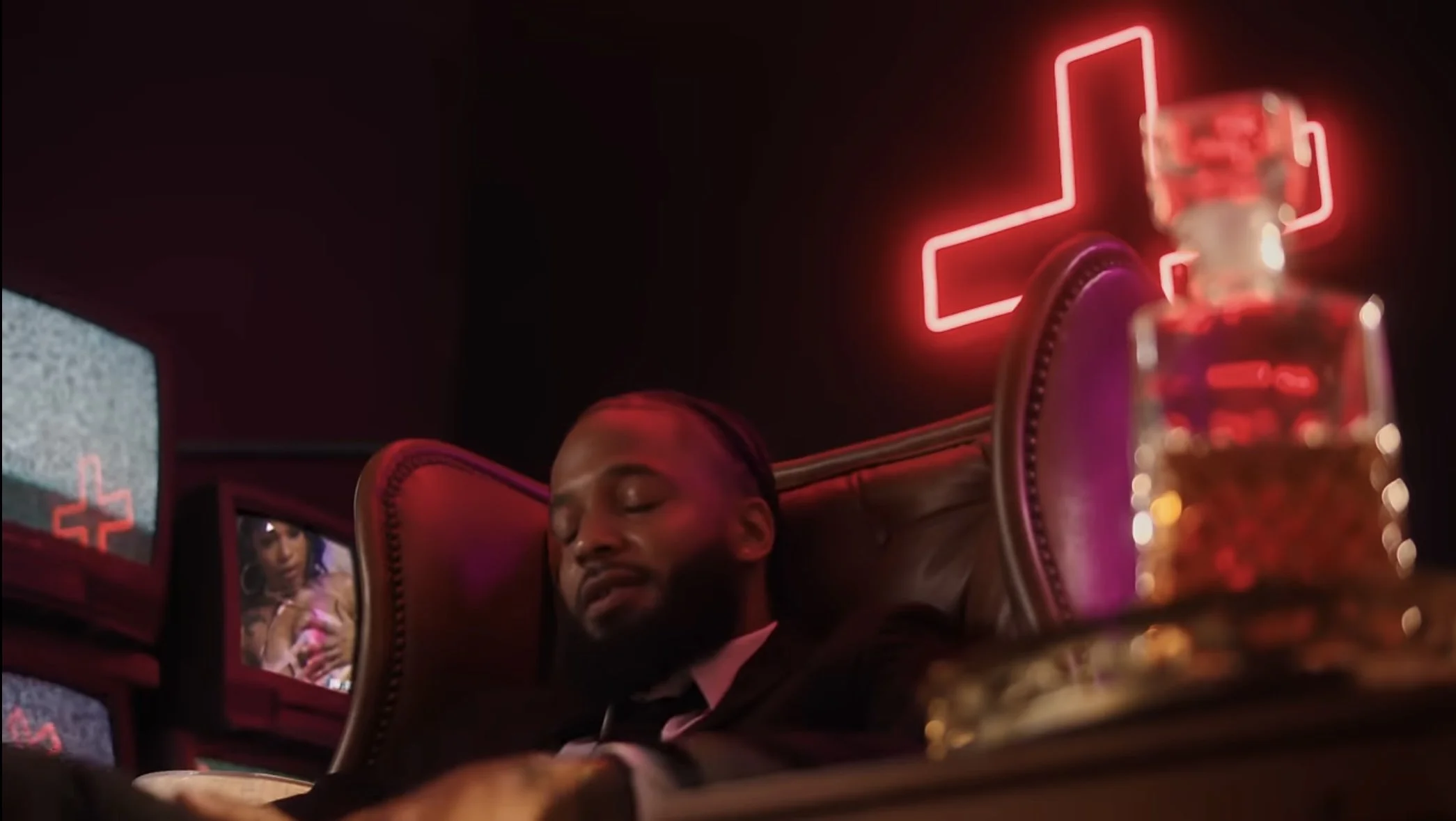 A man with braided hair sleeping at a headrest with a neon red cross sign behind him. There are multiple TV screens nearby, with one displaying static and another showing a woman.
