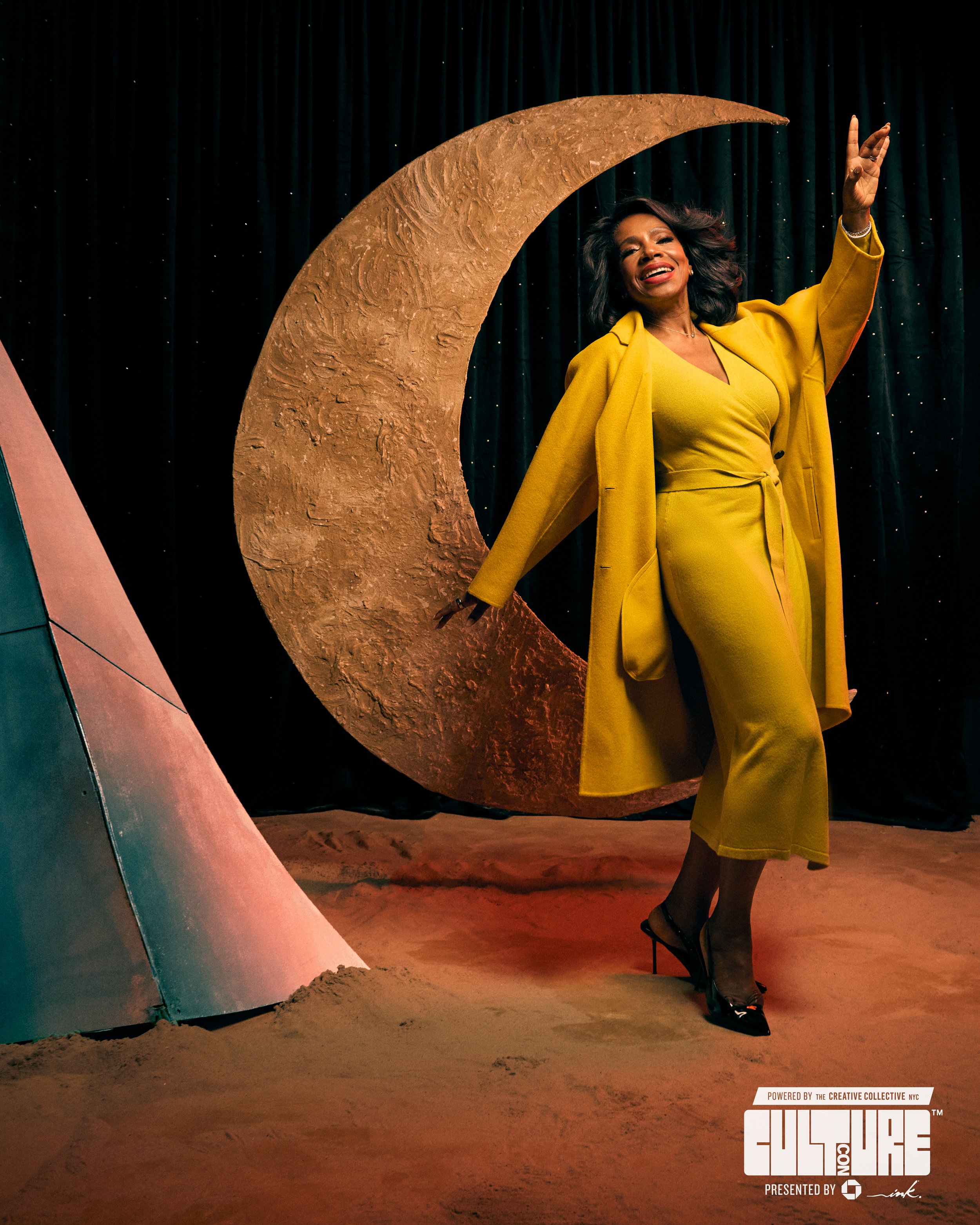A woman in a yellow outfit and coat standing in front of a large crescent moon prop with a starry background. She is posing with a joyful expression.