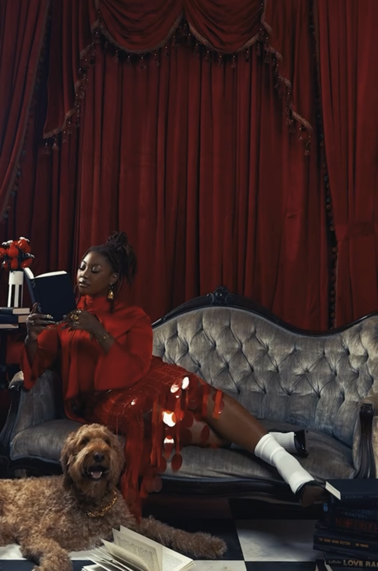 A woman dressed in red and black, lounging on a vintage sofa in front of red curtains, with a dog lying nearby and stacks of books around her.
