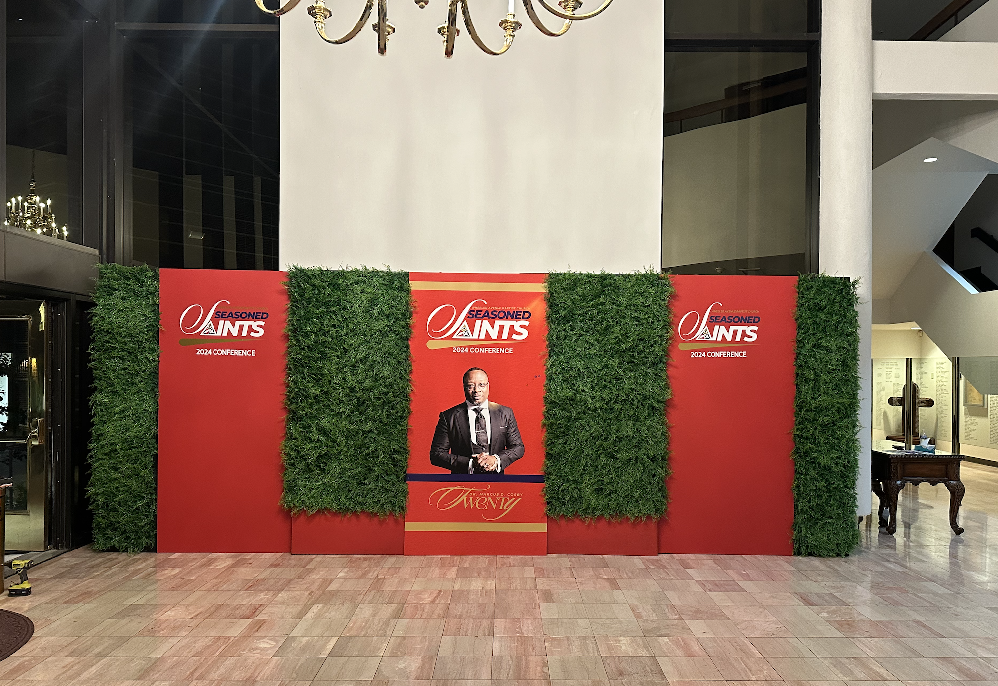 A red step-and-repeat backdrop with green foliage accents and logos for the 2024 Seasoned Saints Conference. In the center, a poster of a man in a suit with the name Dr. Marcus D. Cosby, with the number twenty in stylized font. The setting appears to