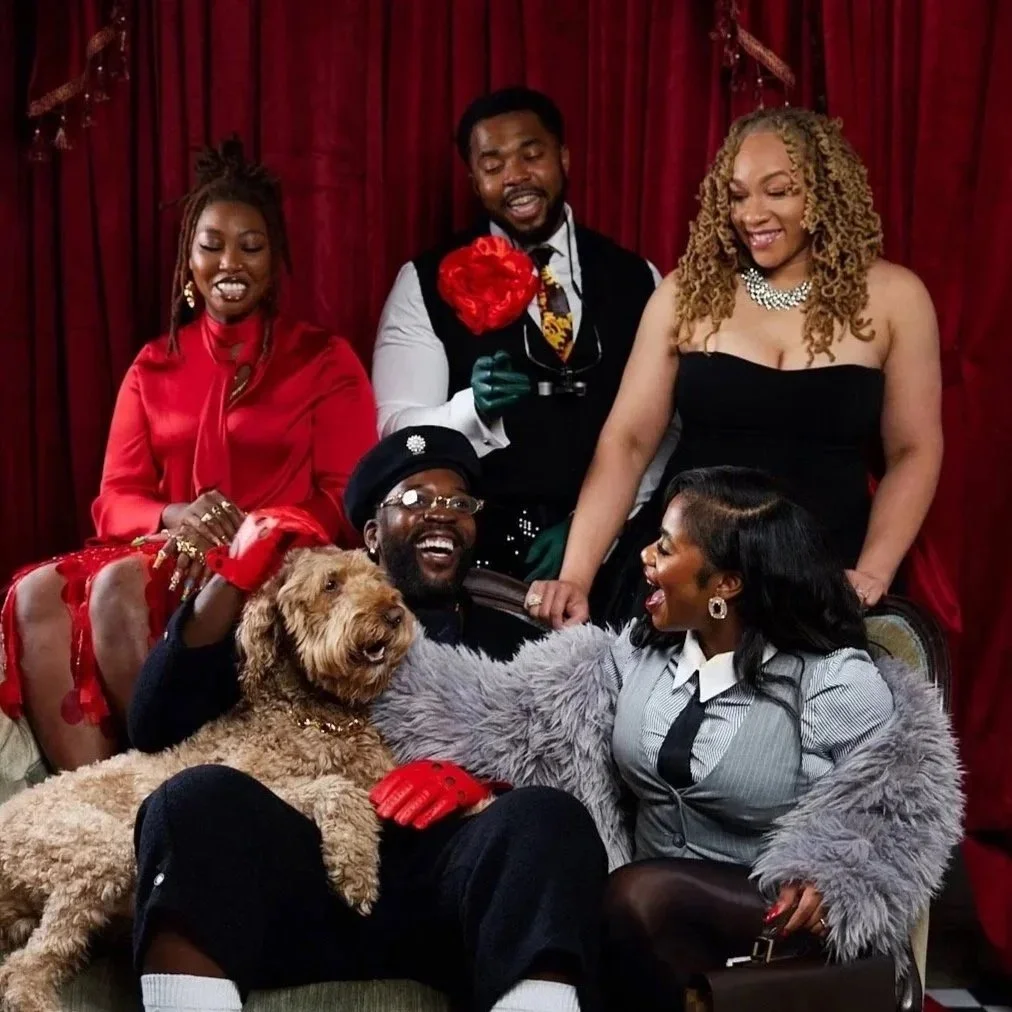 A group of six people and a dog gathered together, smiling and enjoying each other's company in front of a red curtain. They are dressed in elegant and colorful attire, with some wearing accessories like jewelry and gloves.