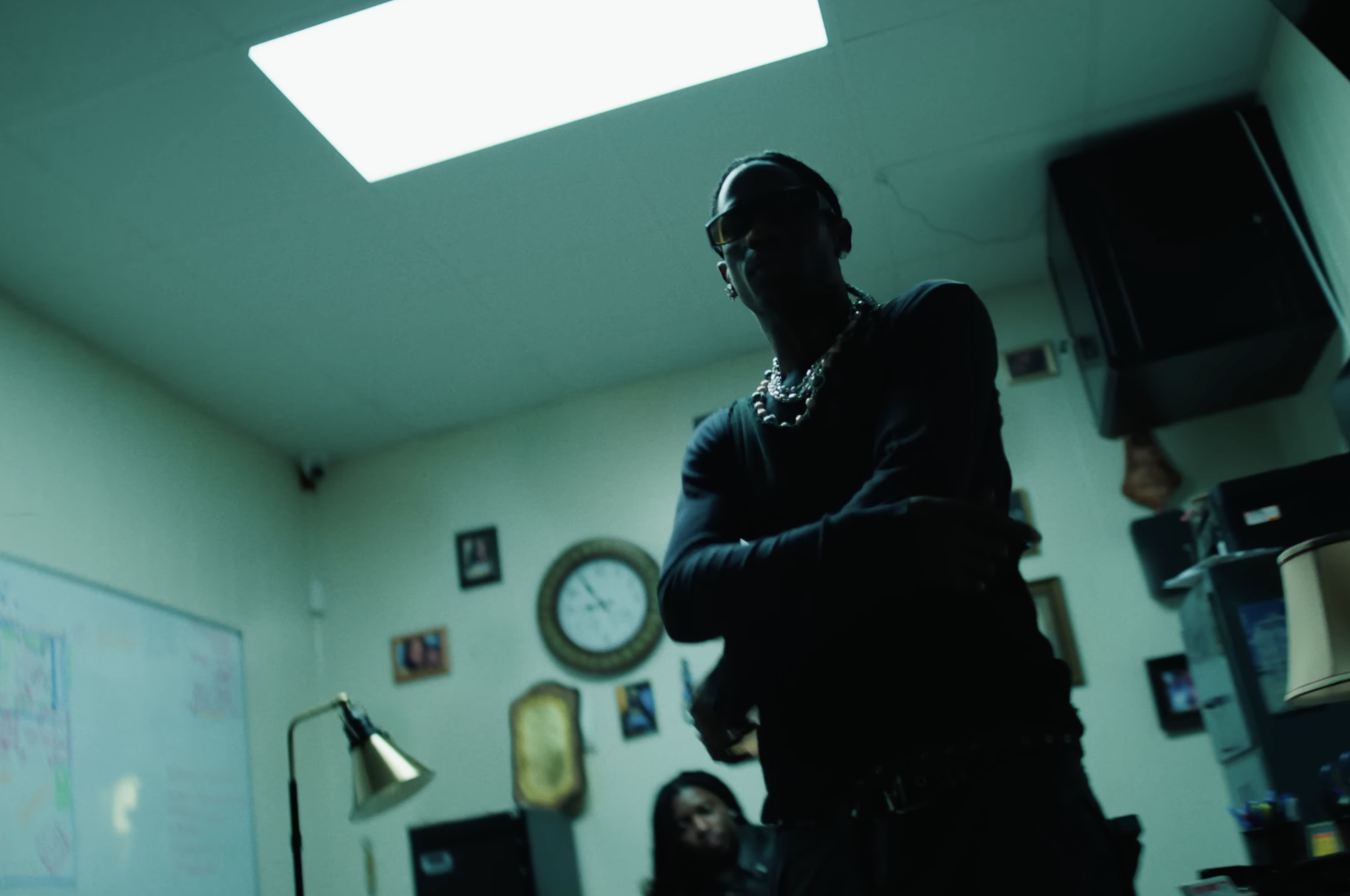 Dark silhouette of a person wearing sunglasses and jewelry in a dimly lit room with a clock and picture frames on the wall.