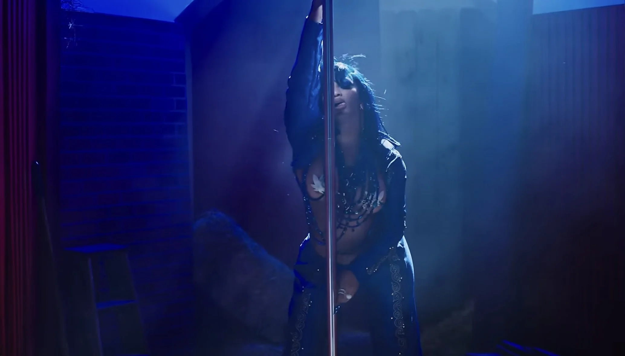 A woman with long dark hair poses with a pole in a dimly lit room with blue and purple lighting, wearing a sheer top and pants with decorative patterns.
