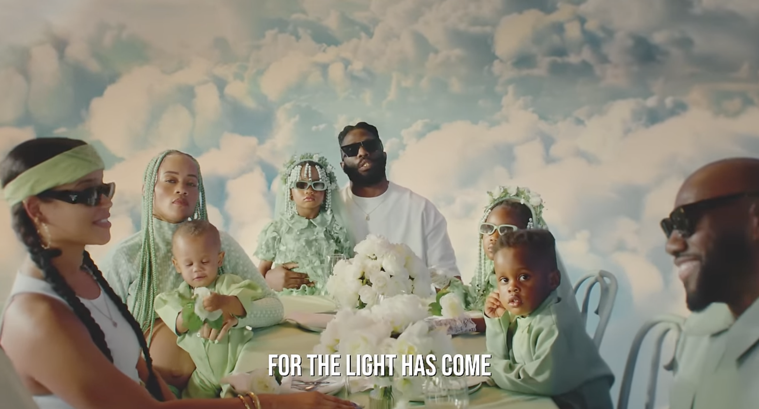 A family sitting at a table with white flowers, wearing green and white clothing, against a sky with clouds, with the text 'For the light has come' at the bottom.