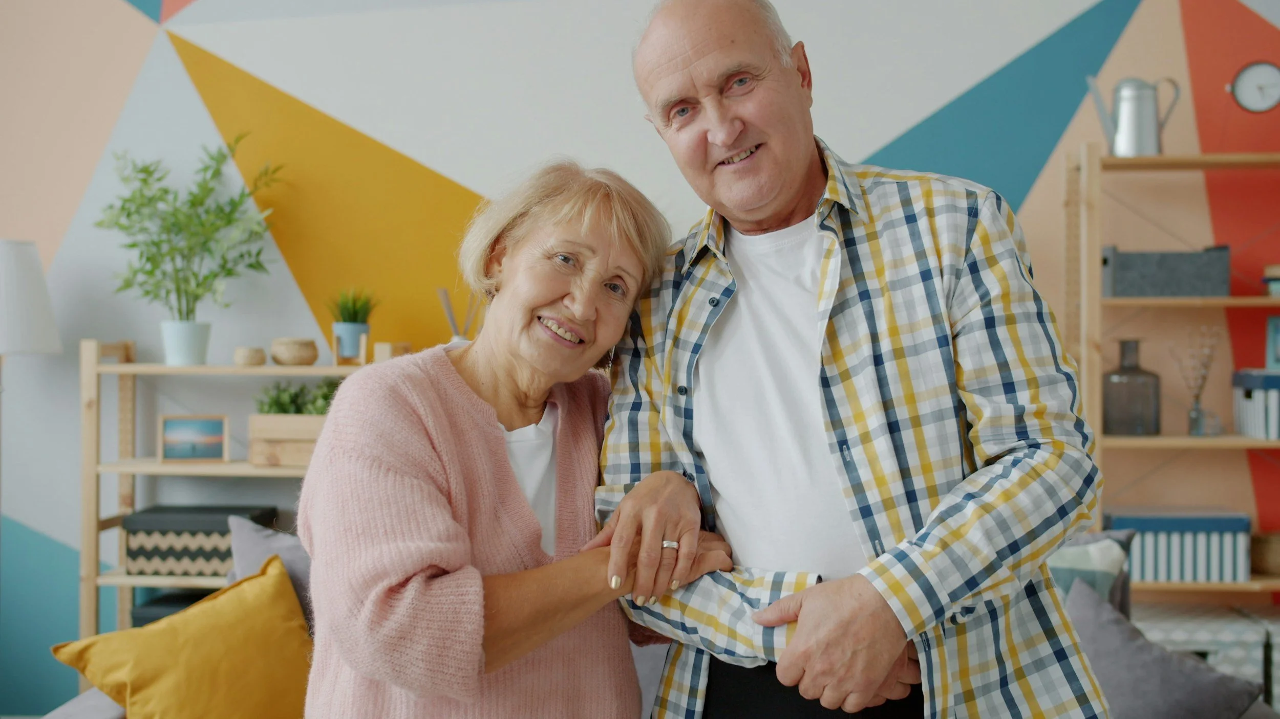 An elderly woman and man smiling and holding hands in a cozy, colorful living room.