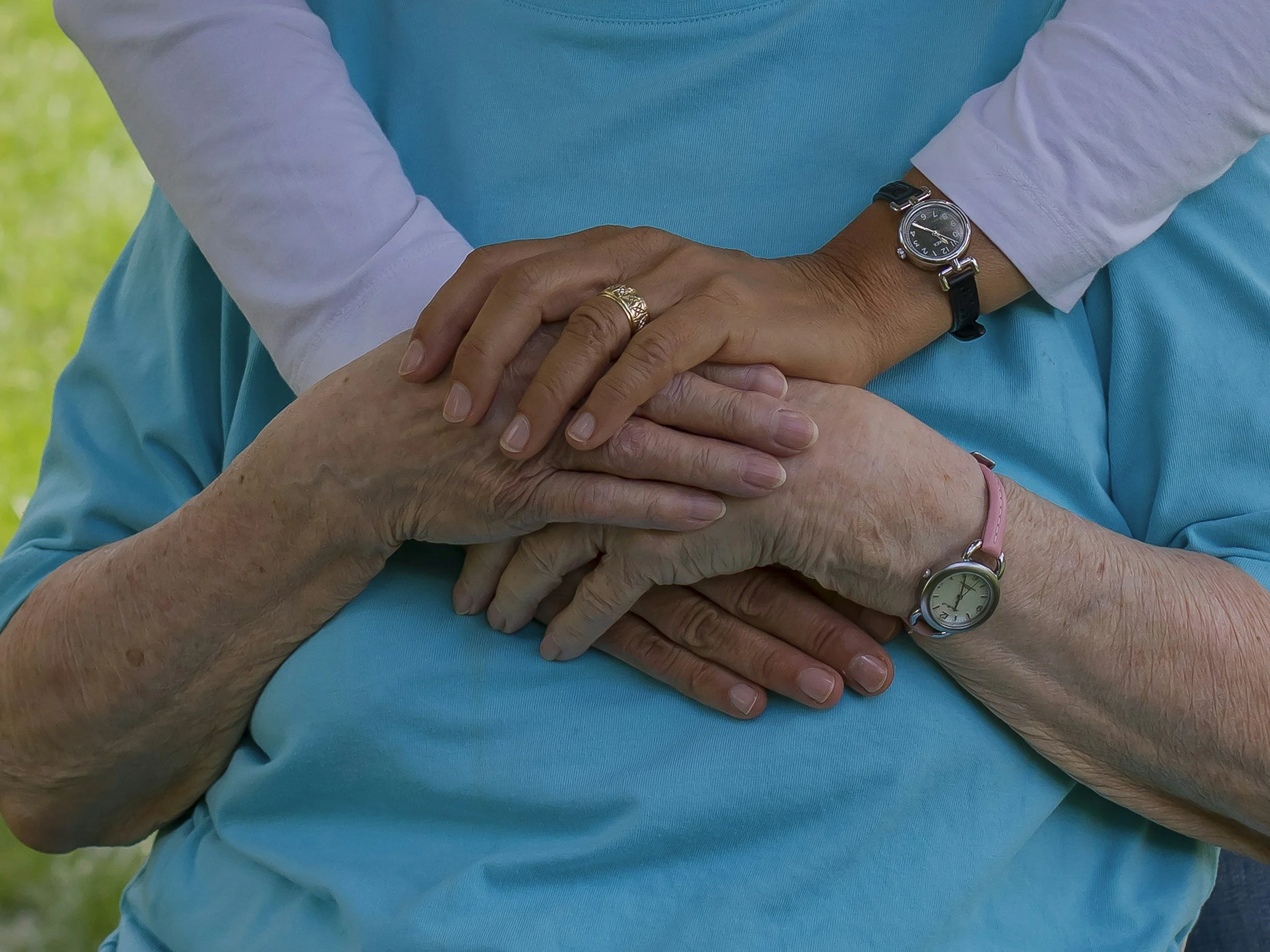 Close-up of multiple hands, from different people, layered on top of each other, placed on a person's stomach. All are wearing watches or rings, with a blurred green outdoor background.