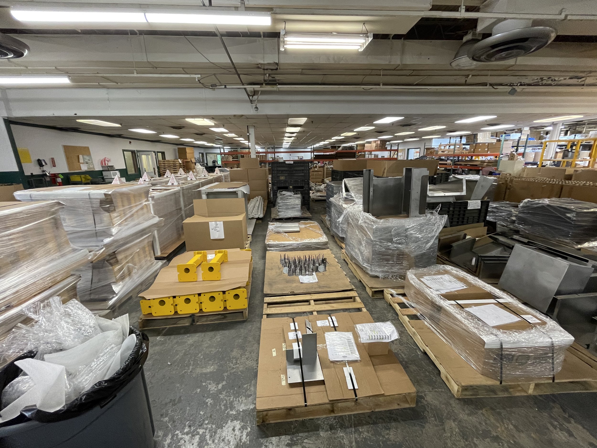 Warehouse or manufacturing facility with metal parts, cardboard boxes, and pallets, some wrapped in plastic, organized on the floor.