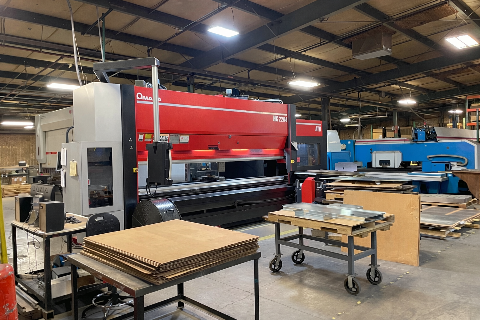 Industrial warehouse with a large red Amada laser cutting machine, stacks of raw wood and metal sheets, and work tables.