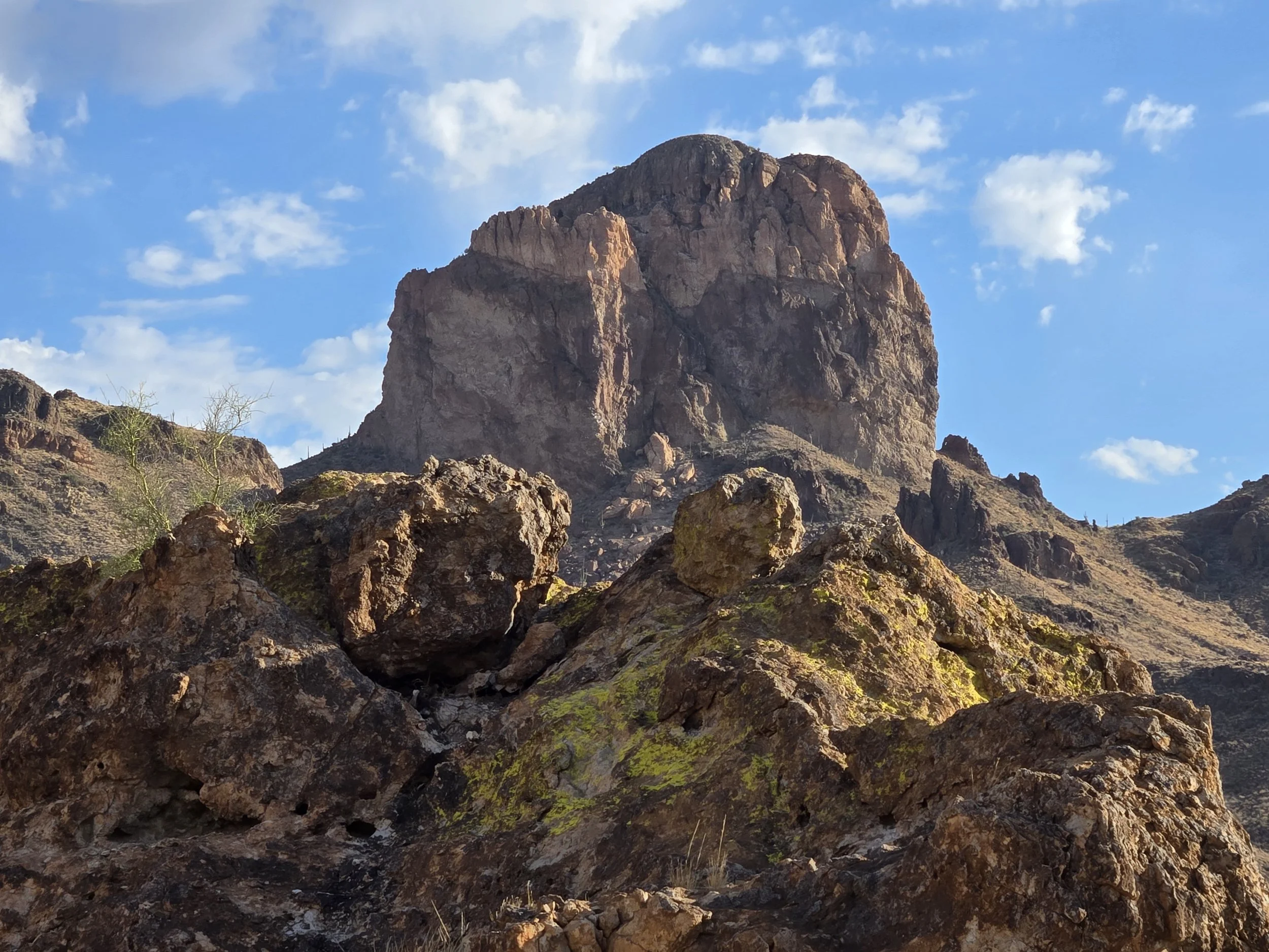 Large rocky mountain with a clear blue sky and scattered clouds in the background. Rocks in the foreground covered with patches of green moss or lichen. Buzzard' Roost, Superstition Mountains, Arizona.