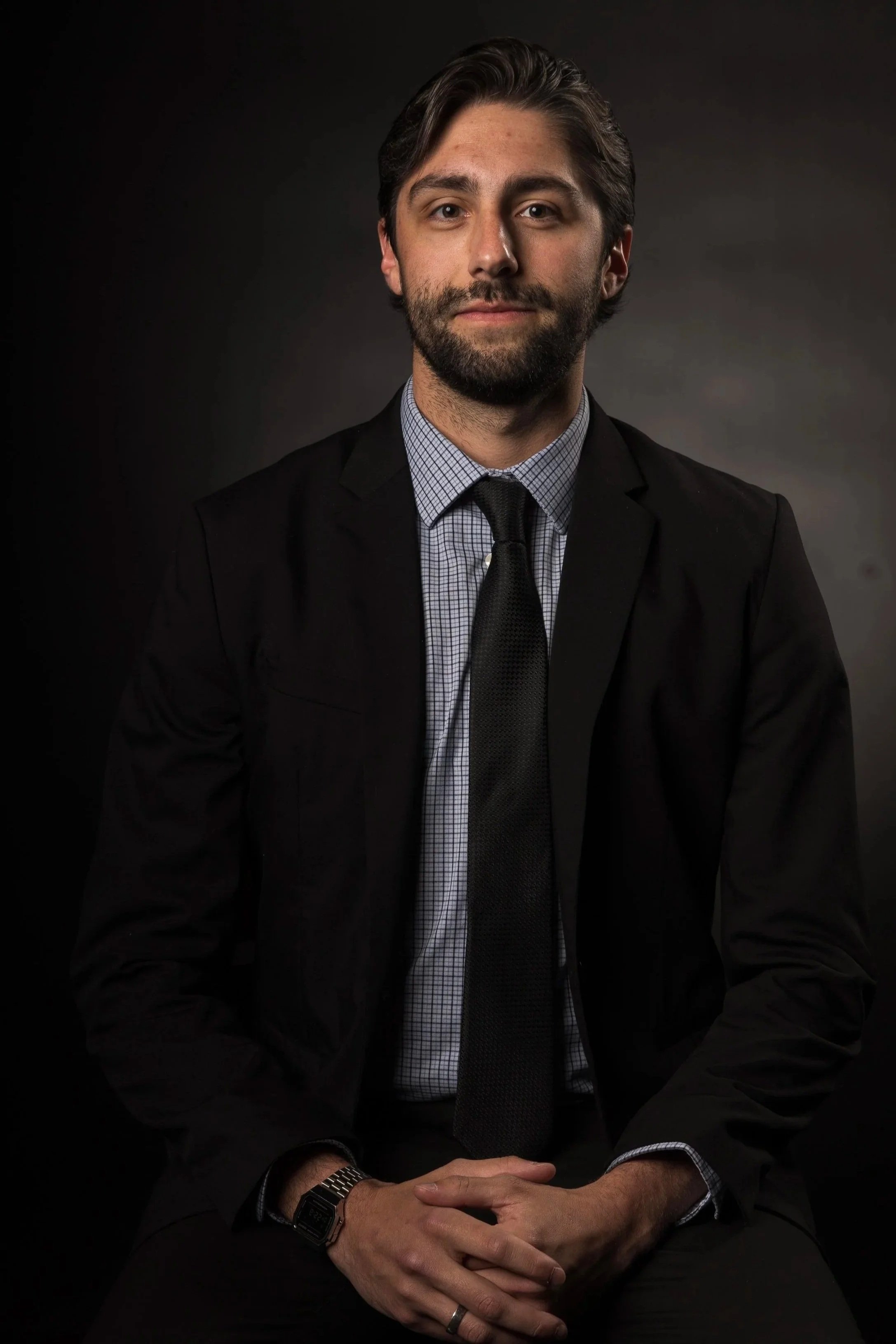 A man with dark hair and a beard, dressed in a black suit, shirt, and tie, sitting with hands clasped, in front of a dark background.