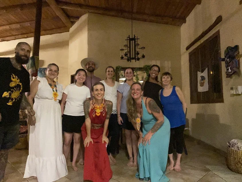 Group of ten people gathered indoors, some wearing casual attire and others in brighter dresses, with a rustic interior featuring wooden ceiling beams and a chandelier.