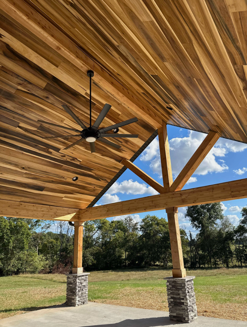 View of underside of a wooden porch ceiling with ceiling fan, supported by stone and wood columns, in a grassy outdoor area with trees and blue sky.