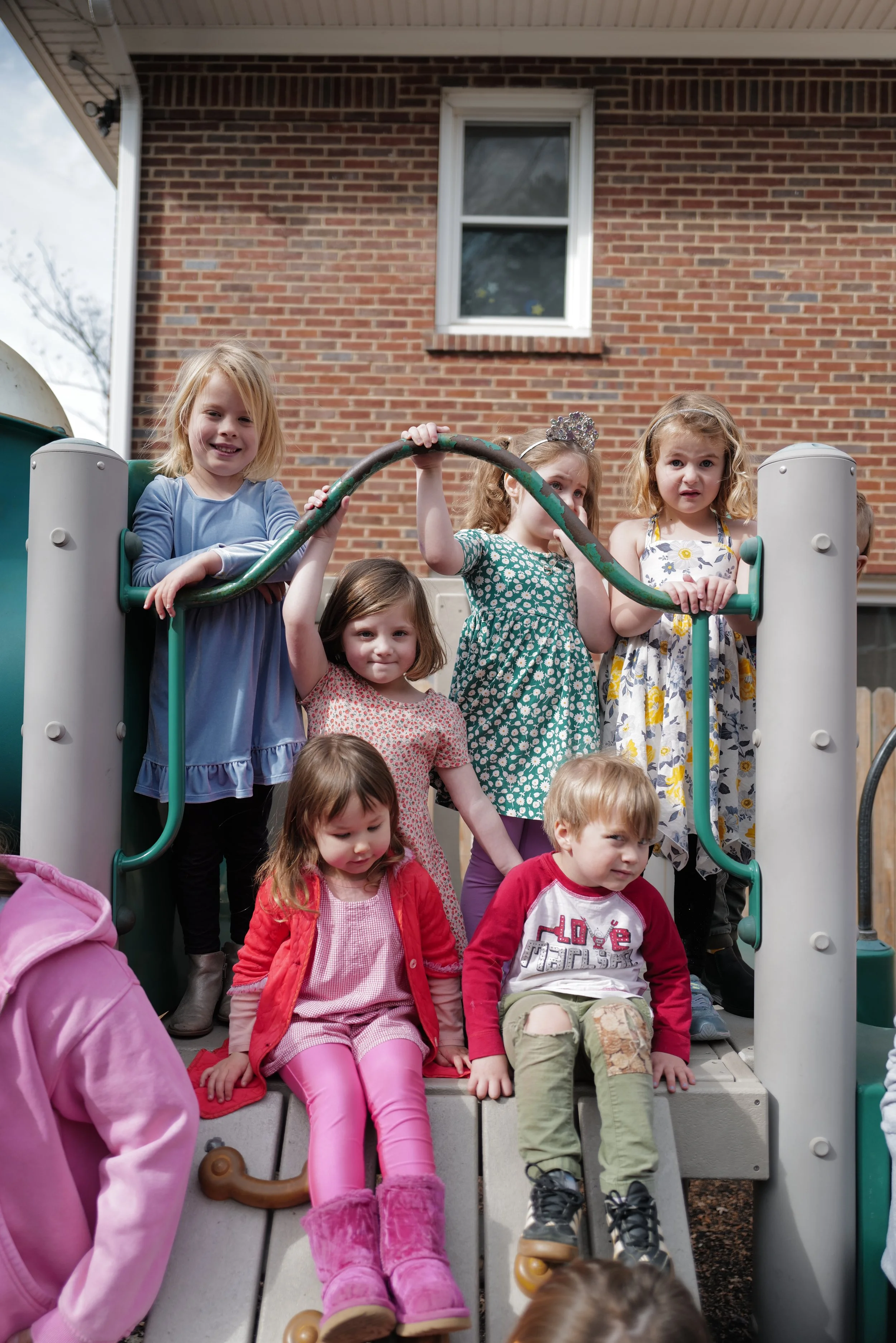 Children playing on a playground structure in front of a brick building, with some children sitting on the steps and others standing on the platform.
