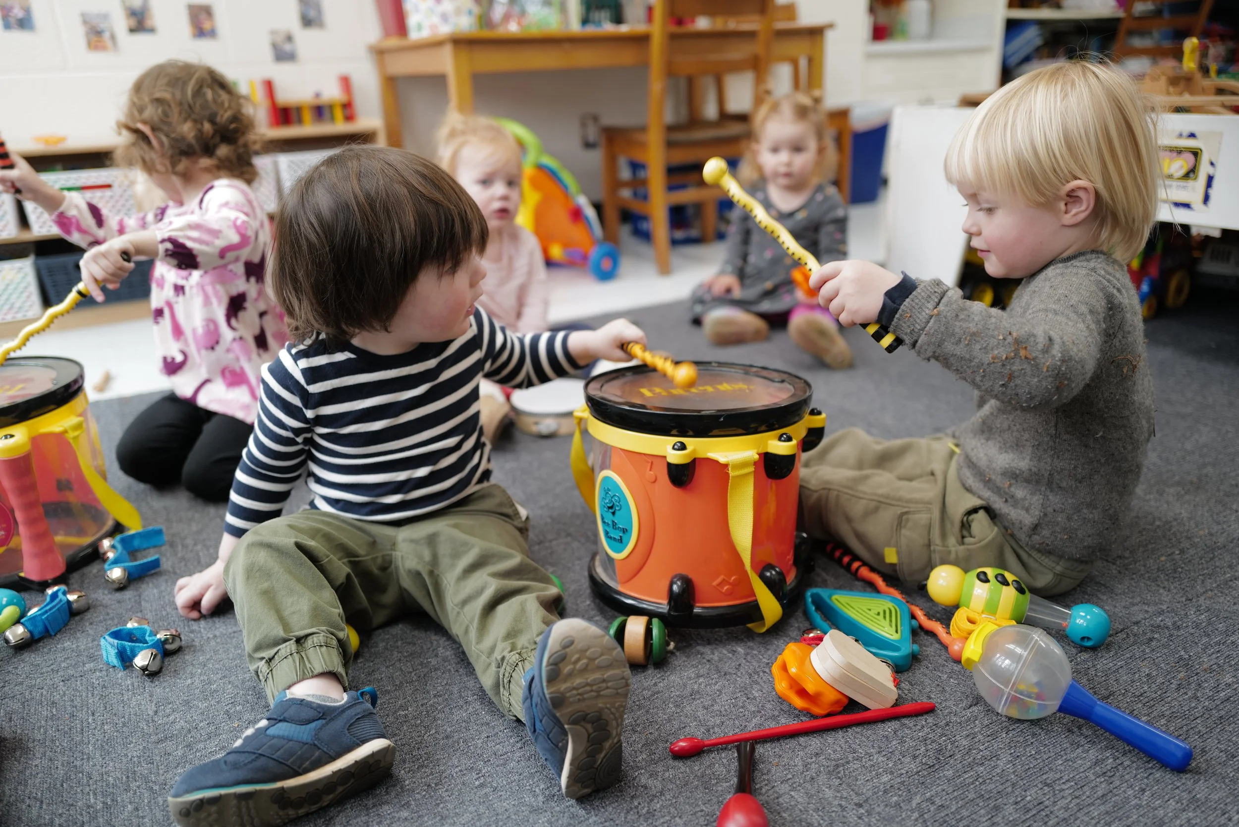 Children in a classroom playing with musical instruments, including drums and other percussion items, sitting on a gray carpet.