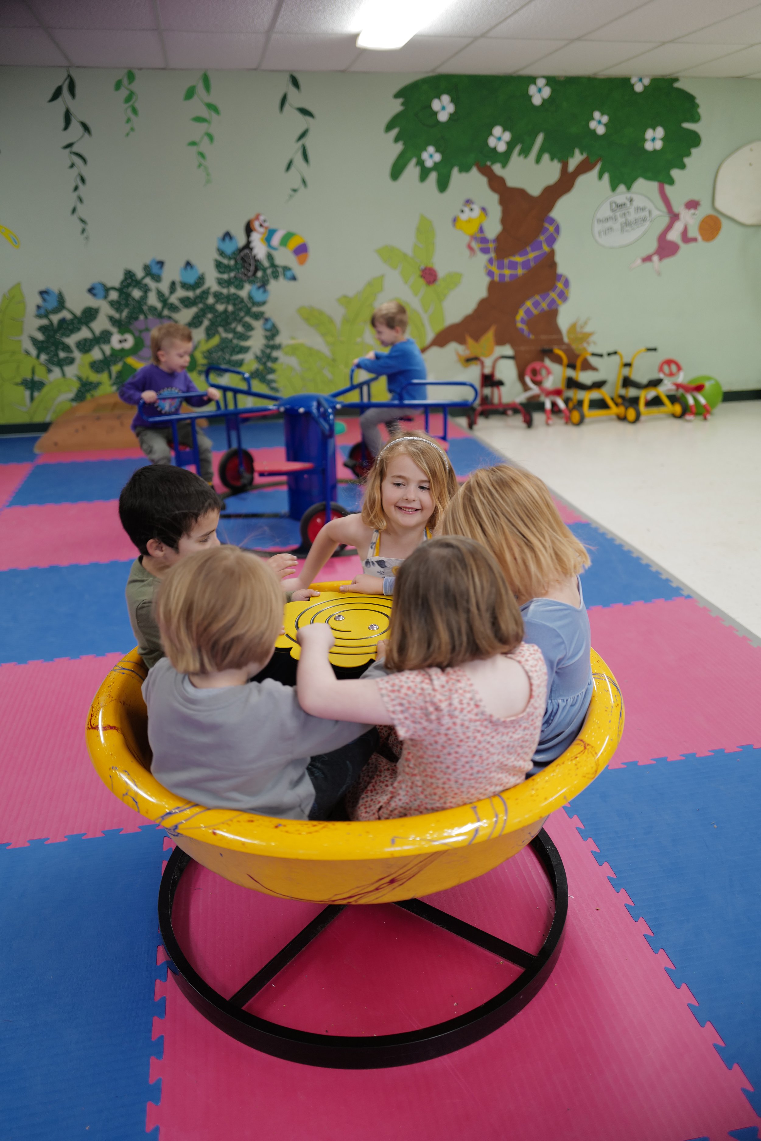 Children playing on a yellow spinning wheel and in the background, children playing in a play area with jungle-themed wall murals.
