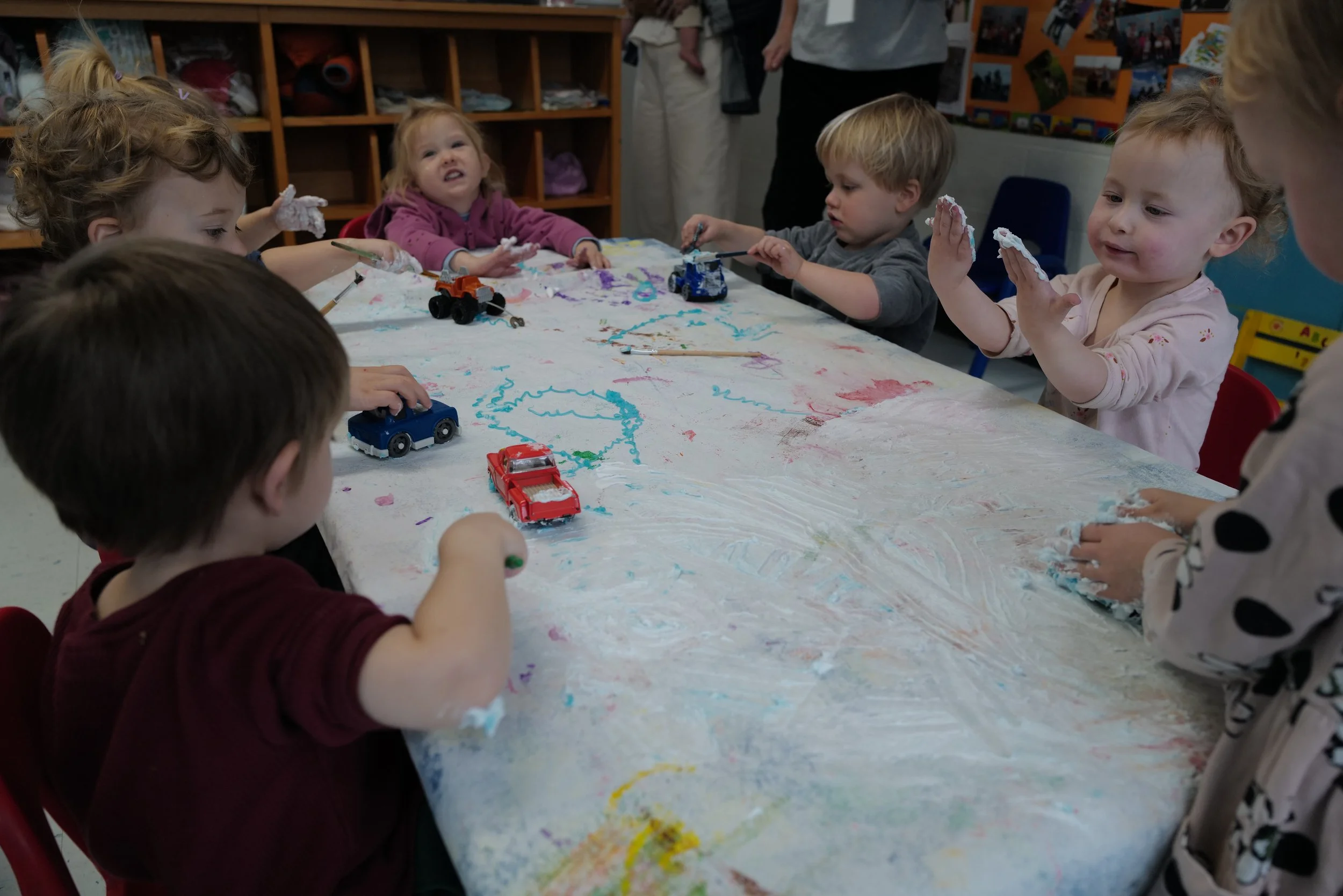 Children engaged in a messy art activity around a large table covered in shaving cream and toys in a classroom.