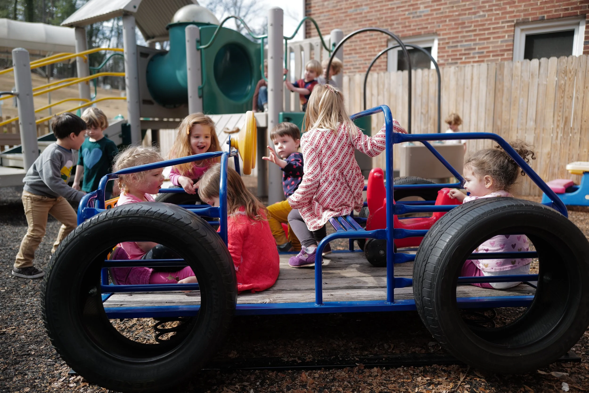 Children playing on a blue wagon in a playground with a wooden fence, slide, and other play structures in the background.
