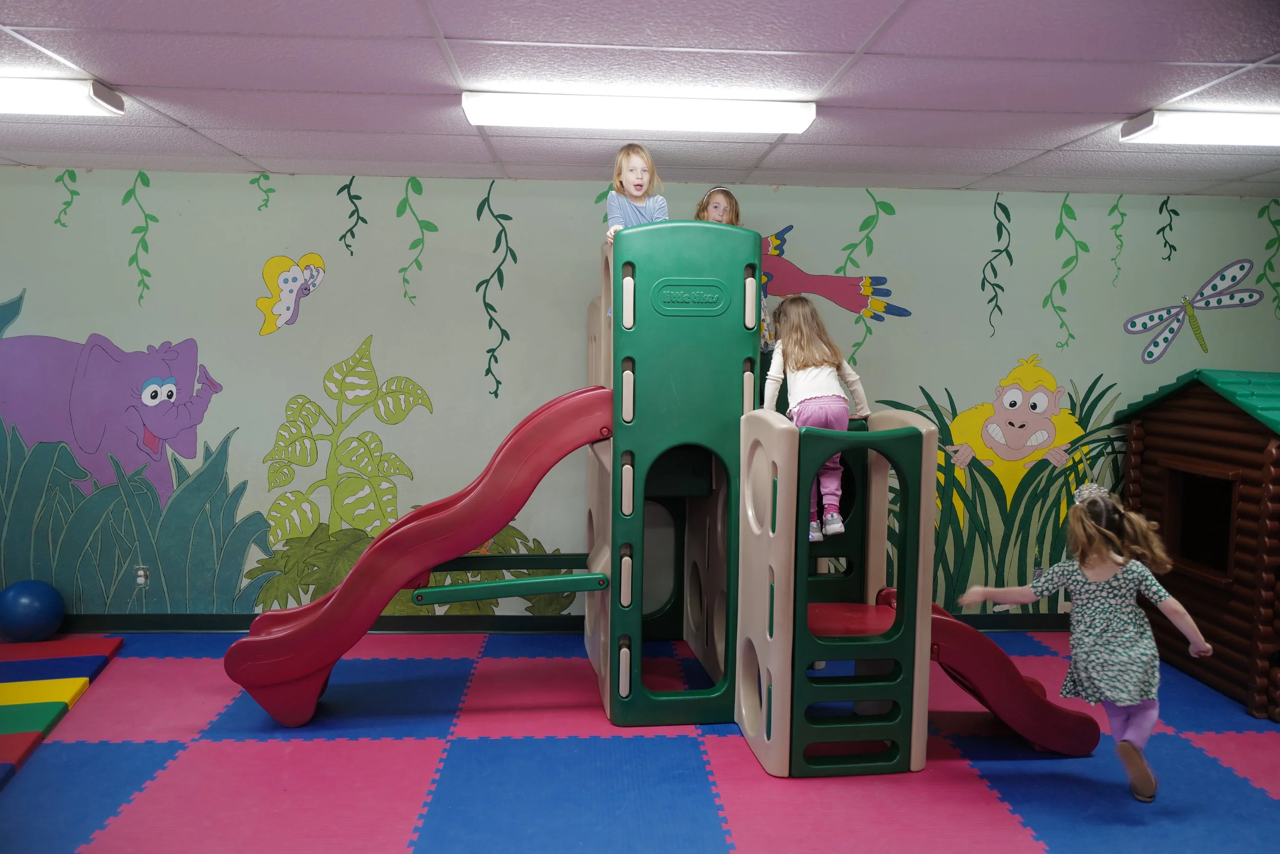 Children playing on a colorful indoor play structure with slides in a children's playroom decorated with jungle and animal mural on the wall.