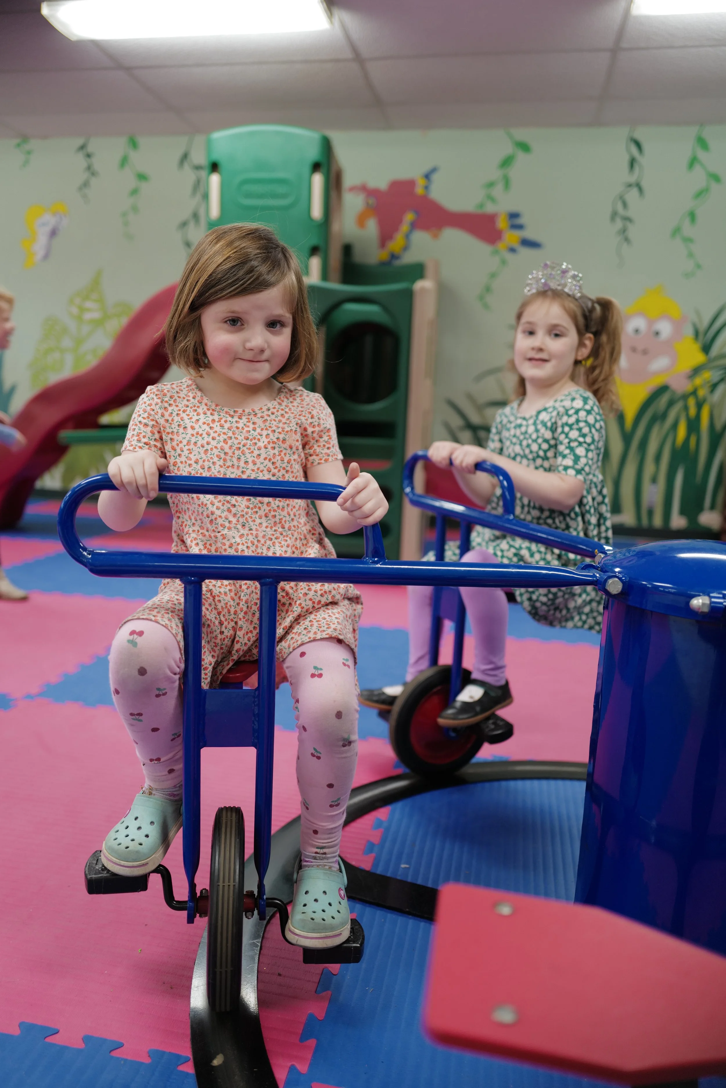 Two young girls playing on indoor playground equipment, one on a blue tricycle and the other on a stationary bike, in a colorful playroom with cartoon character mural walls.