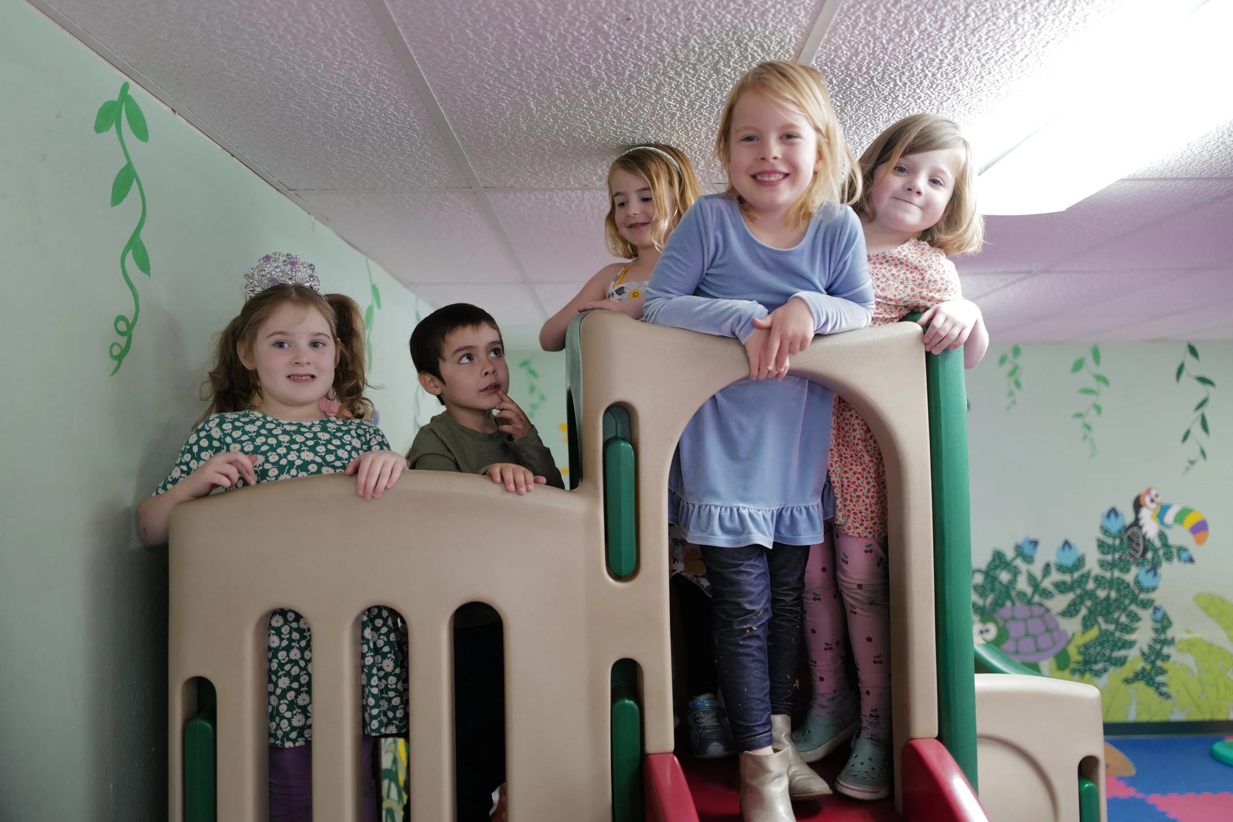 Children playing on a beige and green indoor playground structure with a slide. The background features a painted mural of a turtle and colorful leaves on a light green wall.