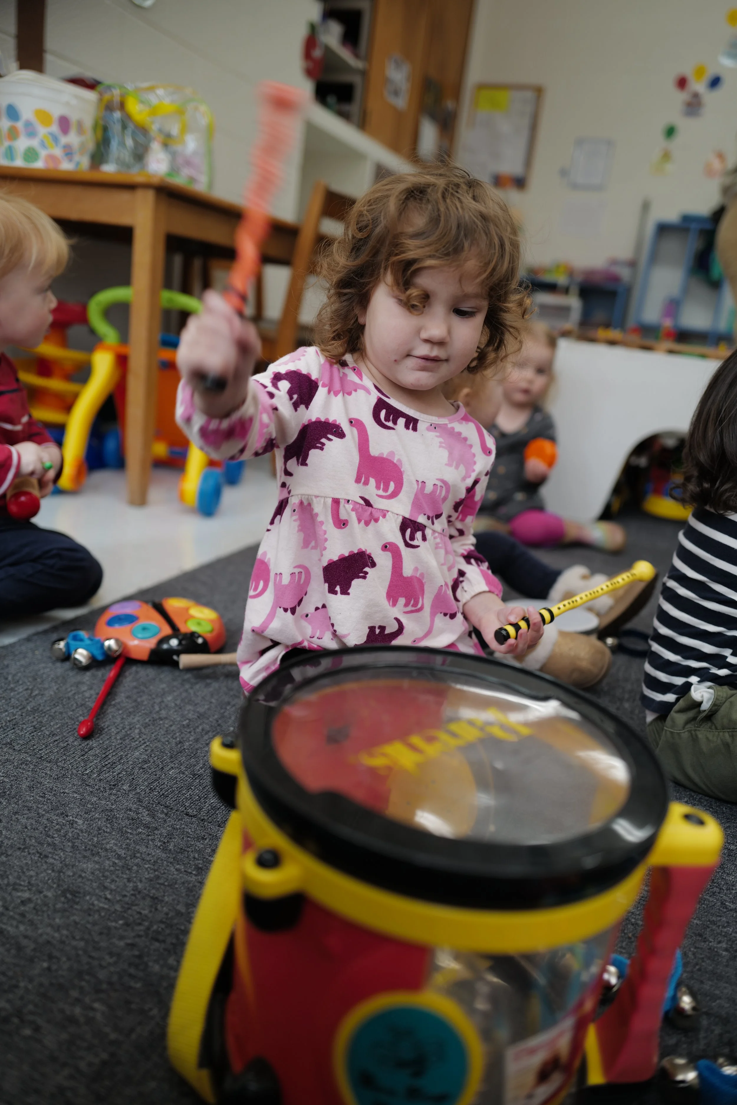 A young girl playing a yellow and red toy drum in a classroom with other children, toys, and furniture in the background.