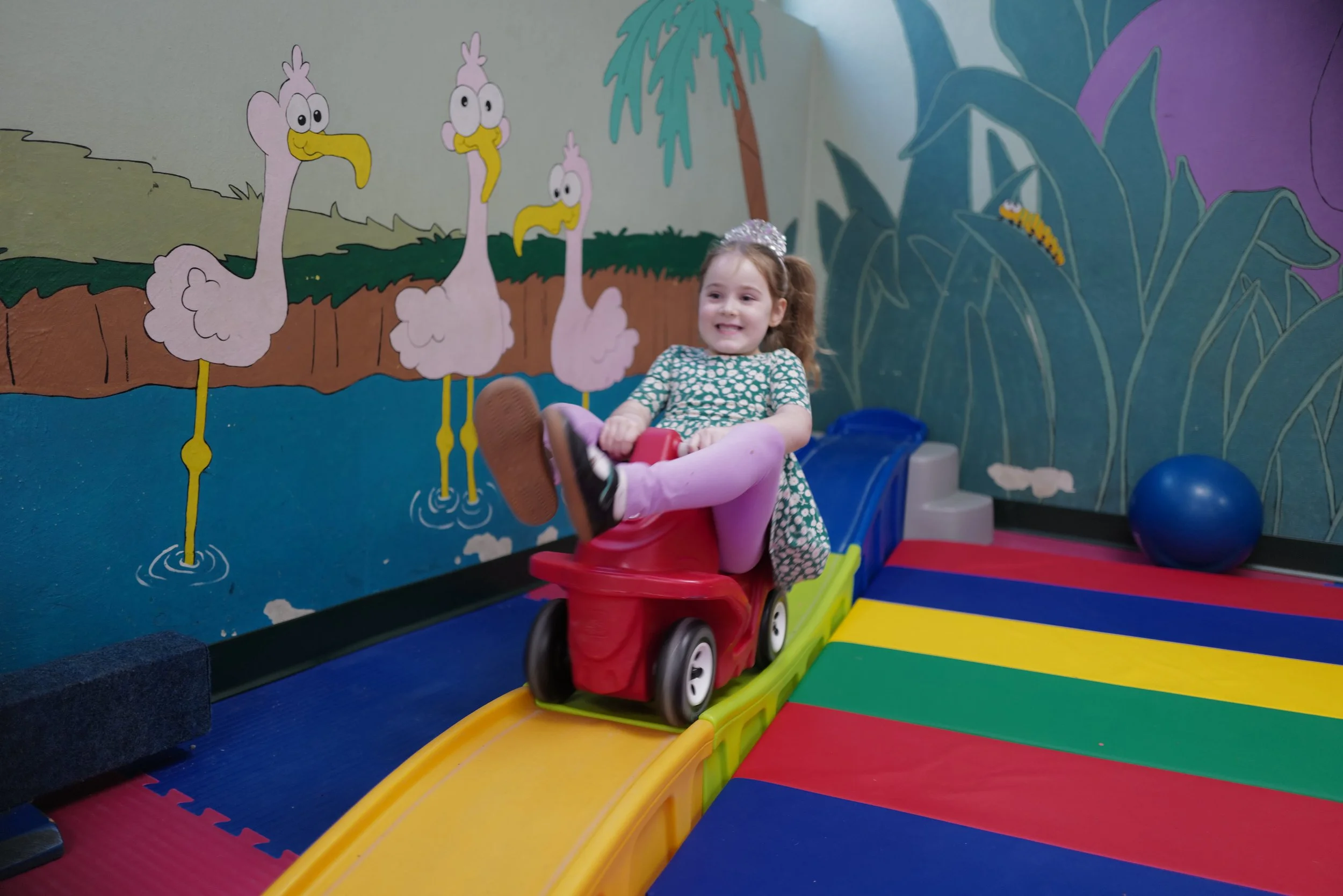 A young girl with a sparkly hair accessory riding a red toy train on an indoor track at a play area with colorful mats and a mural of cartoon flamingos and greenery on the wall.