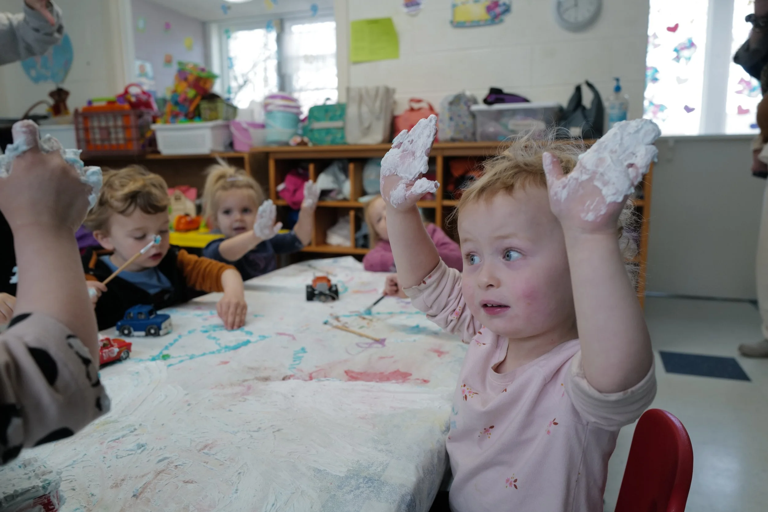 Children sitting around a table during a messy art activity, with some raising their hands covered in shaving cream, in a classroom setting.